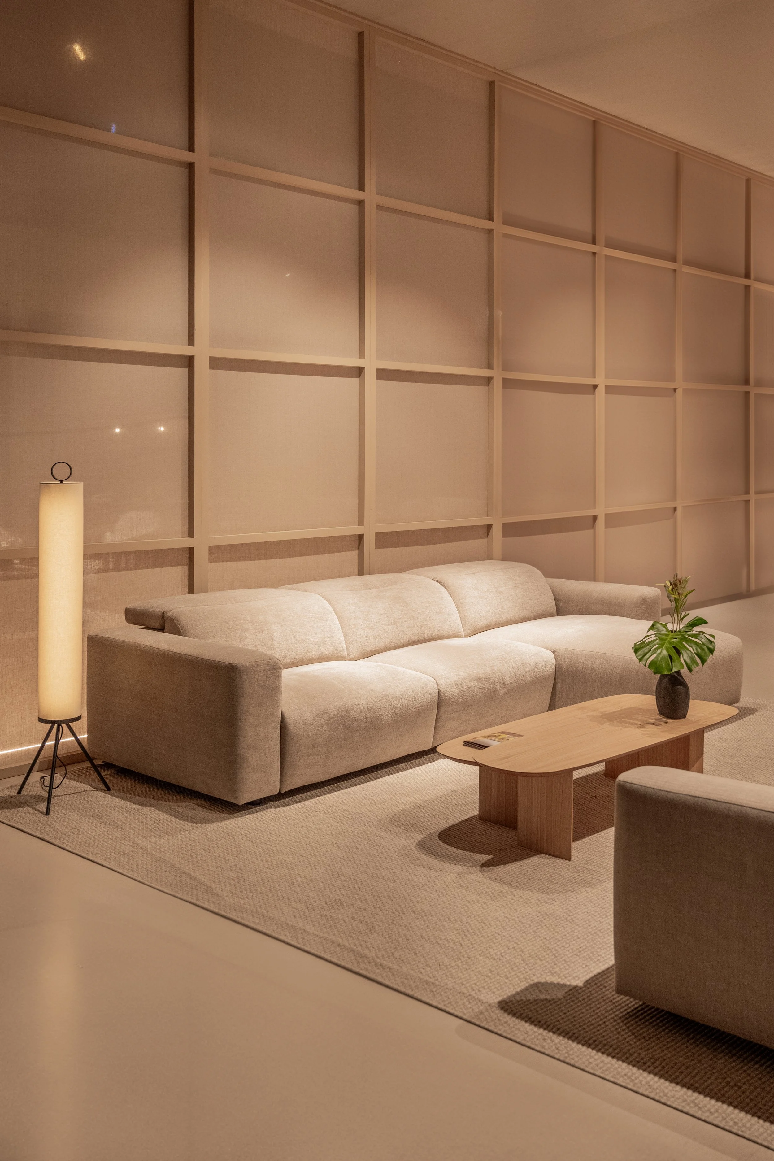 Sala de estar moderna con sofá gris, mesa de madera, alfombra beige, lámpara de pie y planta en una mesa pequeña, pared con divisiones de madera.