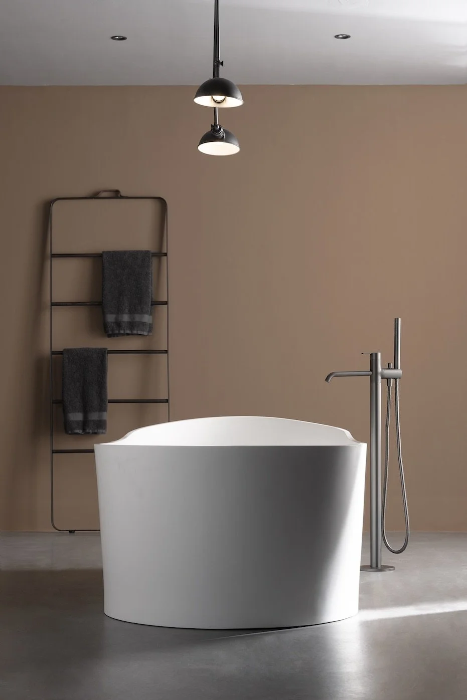 Una bañera blanca de estilo moderno en un baño minimalista con paredes beige, una lámpara negra colgante, y un soporte para toallas negro con toallas grises.