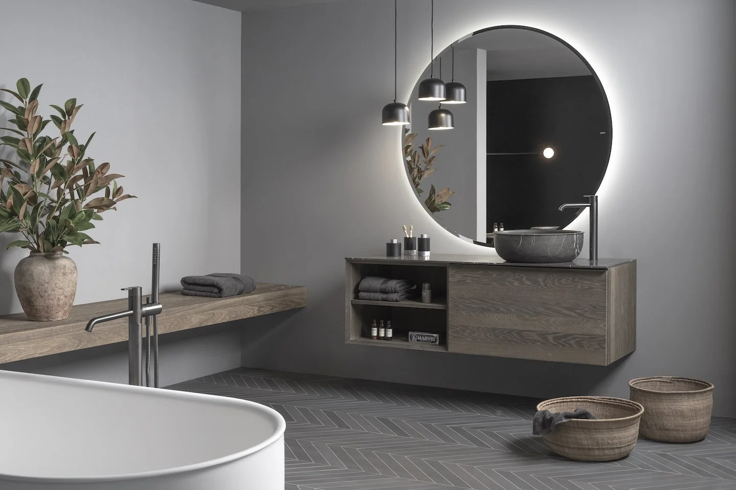 Baño moderno con pared gris, espejo redondo con iluminación LED, lavabo de piedra, y muebles de madera. Hay plantas en cerámicas y cestas de mimbre en el piso, con toallas gris oscuro y accesorios en tonos neutros.