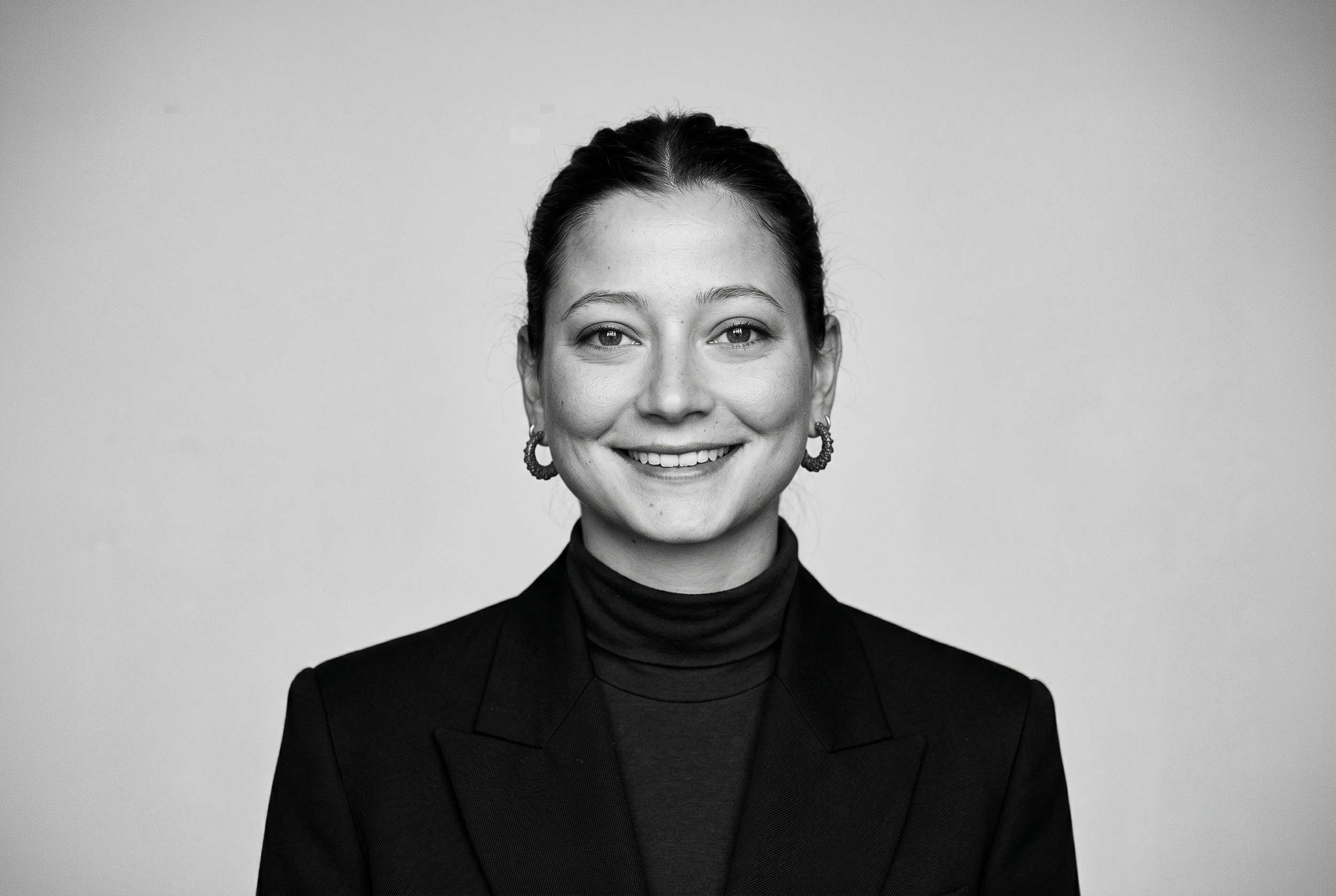 Retrato de una mujer sonriendo, con cabello oscuro recogido, usando aretes grandes y ropa negra. Fondo sencillo y claro.