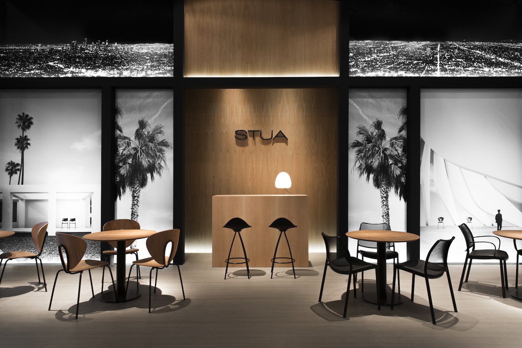Interiores de un espacio moderno con mesas y sillas de madera y negras, paredes con fotografías en blanco y negro de palmeras y la ciudad, un fondo de madera con letras que dicen 'STUA' y una pequeña mesa con una lámpara