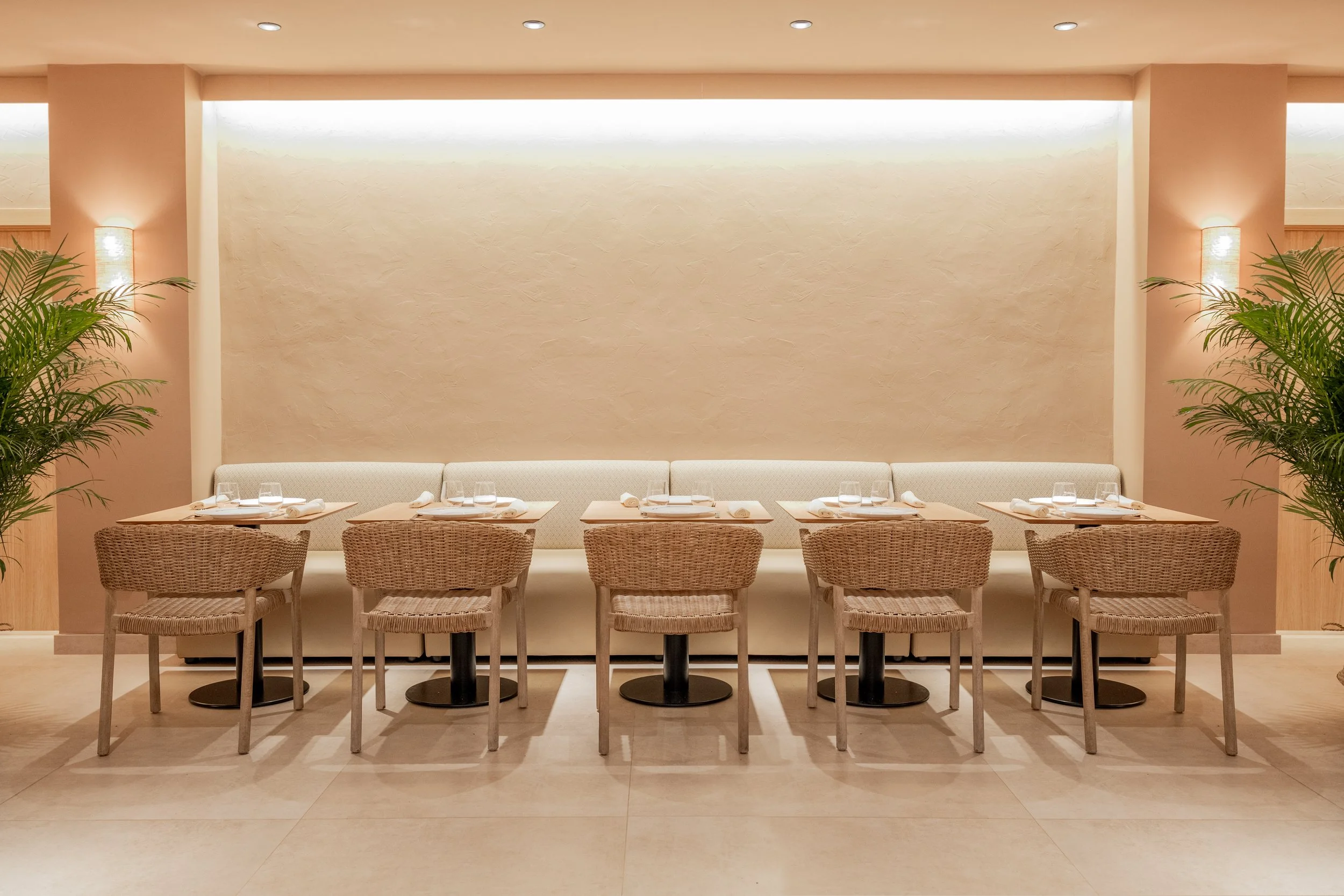 Un comedor con una larga mesa y sillas de mimbre, paredes en tono naranja claro, iluminación suave y plantas a los lados.