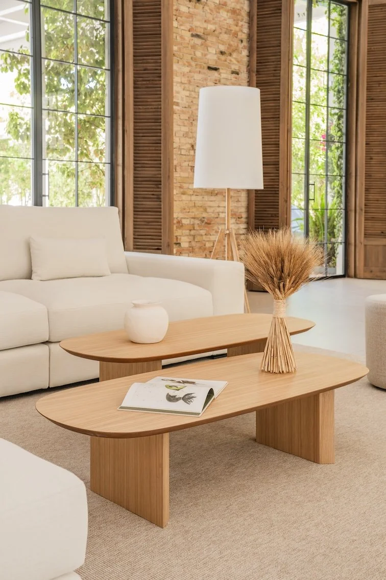 Salón con sillón blanco, mesa de madera, lámpara de pie, florero con trigo de color marrón, paredes de ladrillo y ventanas grandes con vista a naturaleza.
