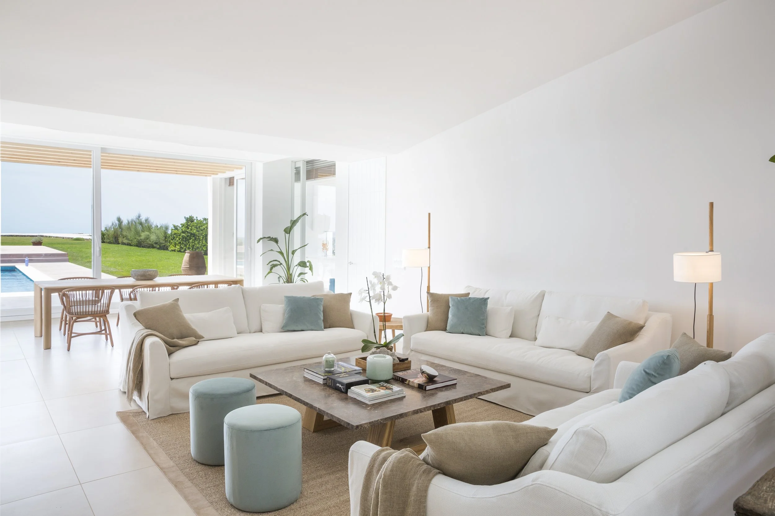 Sala organizada con sillones blancos, cojines en tonos pastel, mesa de centro con libros y velas, plantas verdes y ventanas grandes con vista al exterior con césped y piscina.