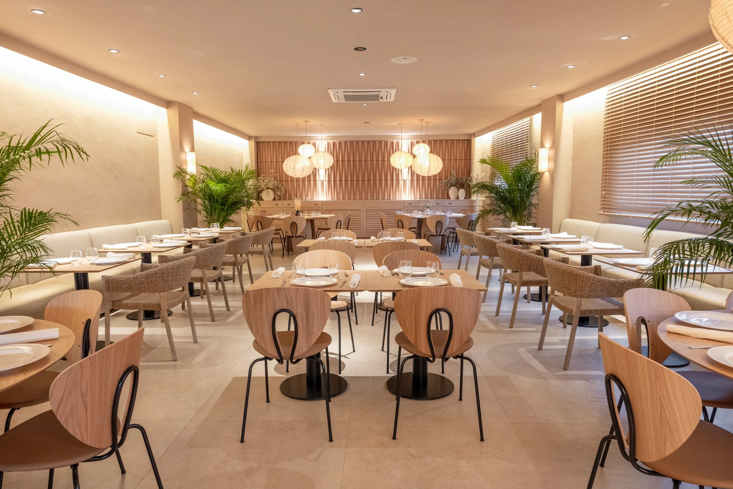 Interior de un restaurante con mesas y sillas de madera, decorado con plantas verdes y luminarias de papel.
