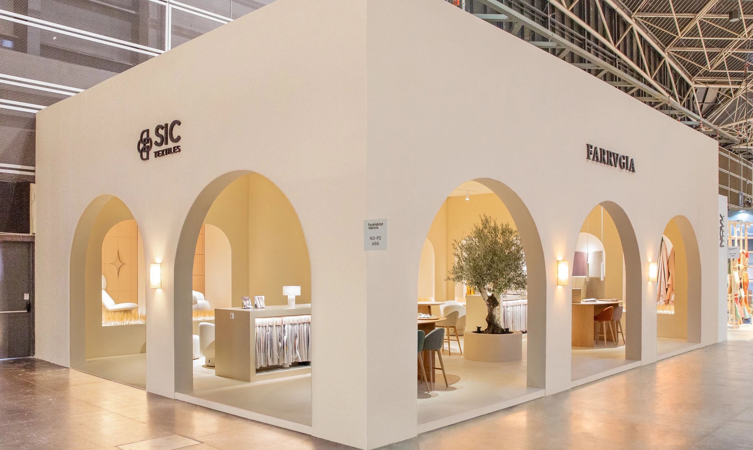 Recinto de exhibición con paredes blancas, arcos y mobiliario moderno en color claro, con plantas decorativas y señalización de marcas como SIC Textiles y Farrugia.
