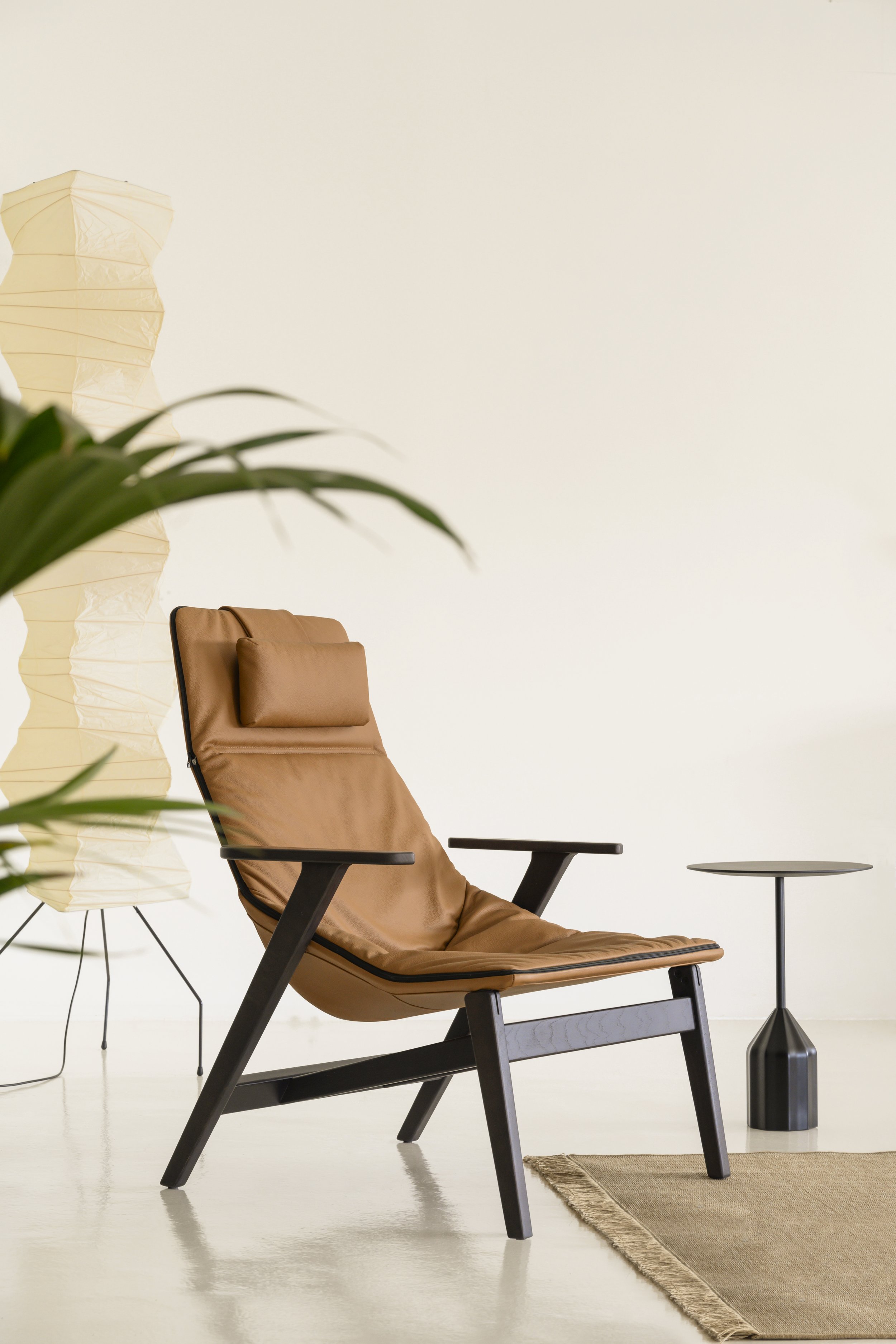 Silla de sala moderna de madera con asiento y respaldo de cuero marrón y cojín, mesa lateral negra, lámpara de piso blanca y lámpara de mesa negra, en una habitación con suelo de madera y pared clara con alfombra.