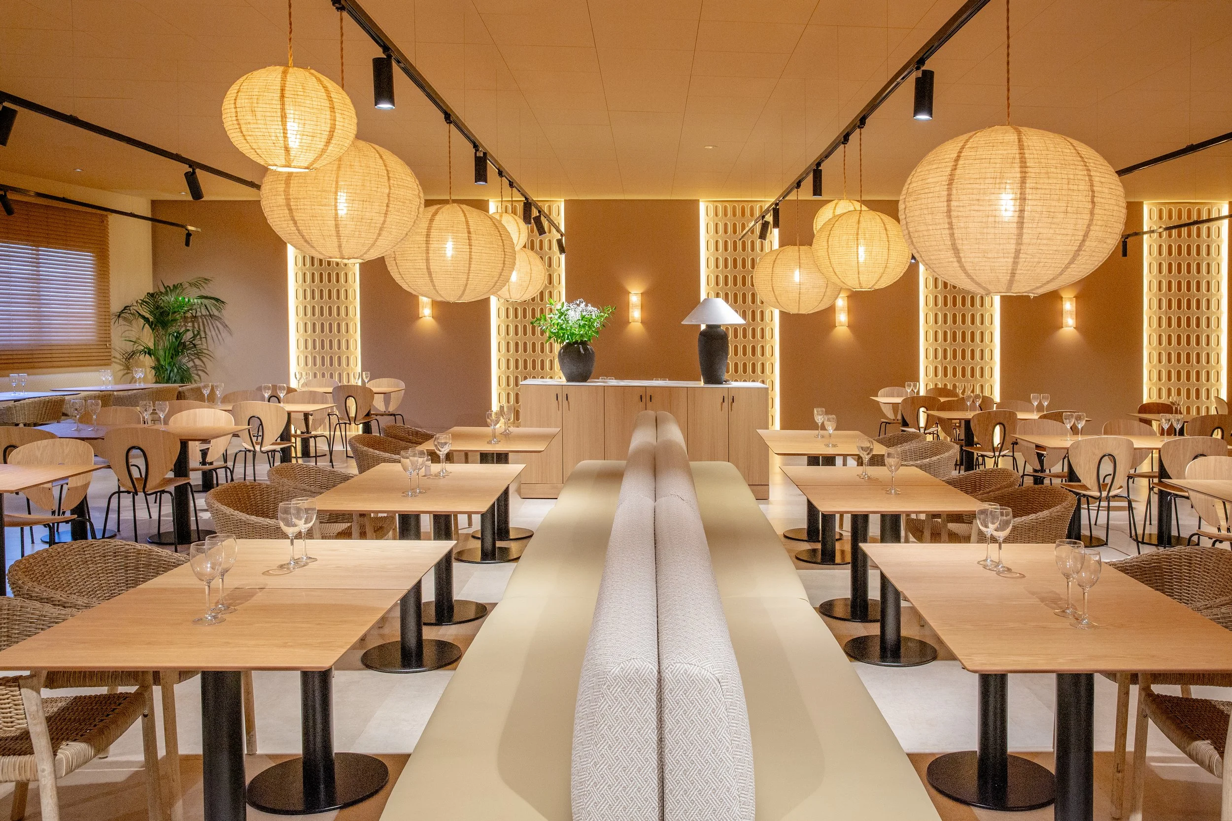 Interior de un restaurante con mesas de madera, sillas de mimbre, lámparas de papel en el techo, y paredes decoradas con luces empotradas y plantas.