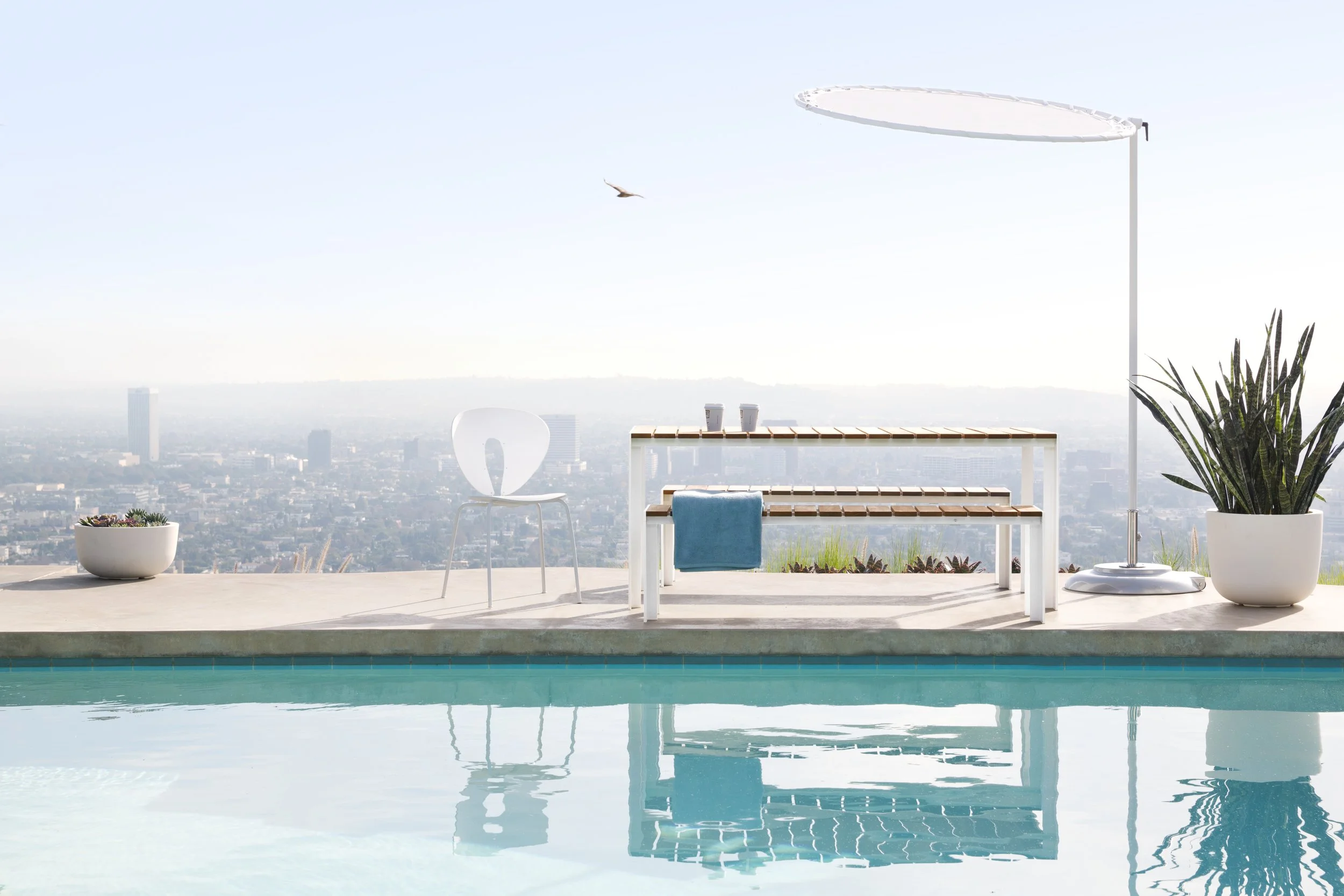 Vista de una piscina en un espacio abierto con muebles blancos, plantas en macetas grandes, un sombrero de sol y una vista panorámica de la ciudad en el fondo, con cielo despejado y un ave en vuelo.