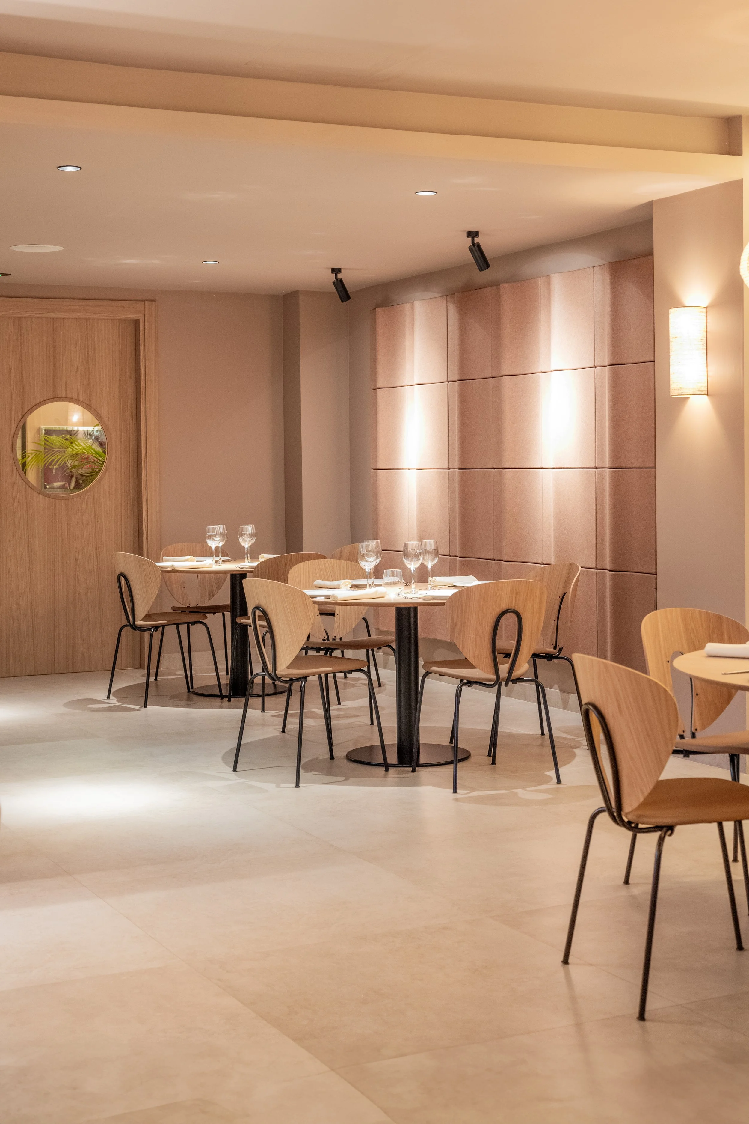 Sala de restaurante con mesas y sillas de madera clara, decorada con paredes beige y una pared tapizada rosa, iluminación suave y lámparas de pared.