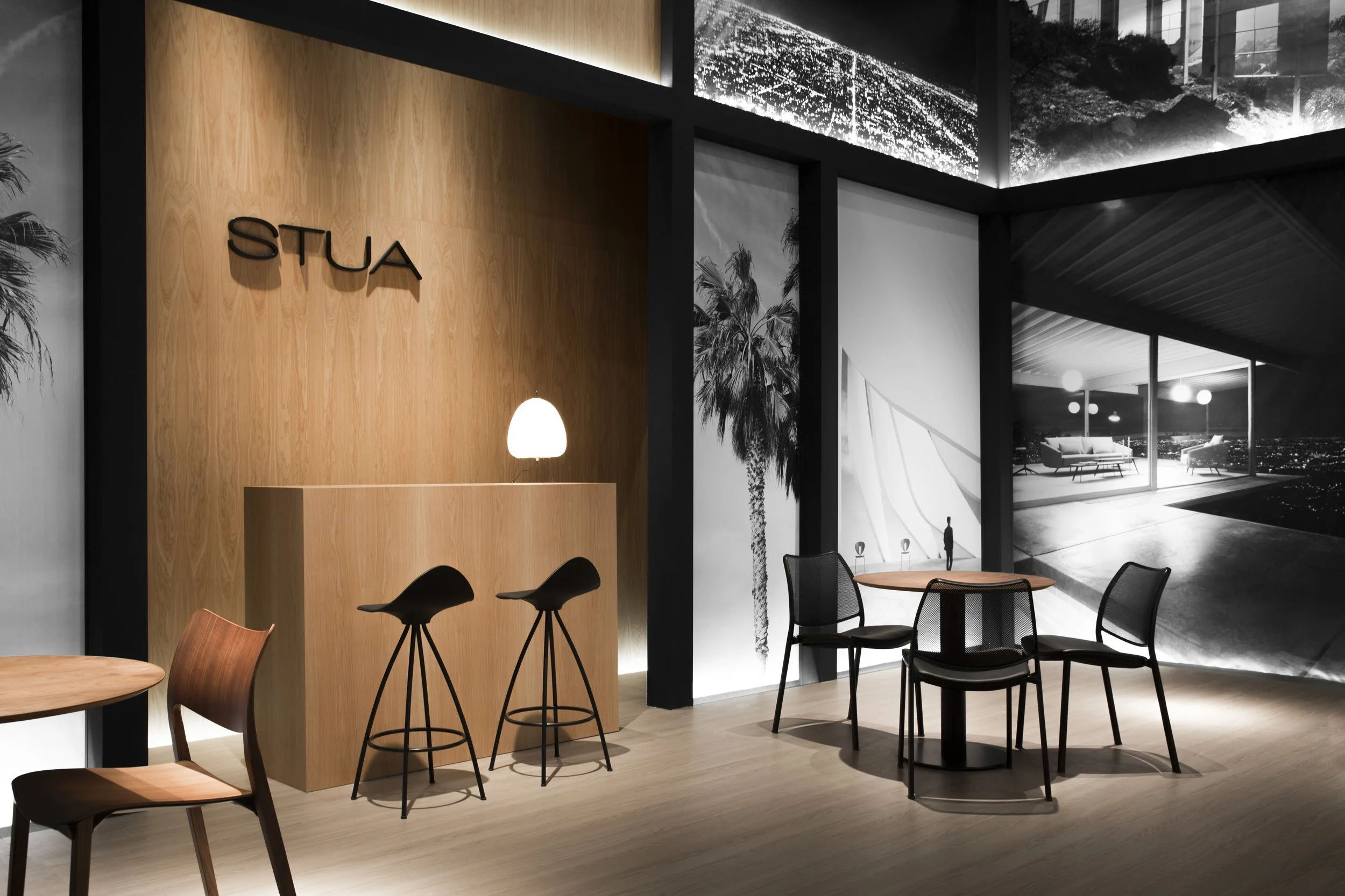 Interiores de un espacio moderno con paredes de madera y ventanas grandes que muestran una vista de la ciudad con palmeras. Hay una mesa redonda con sillas y una pared con el letrero 'STUA'.