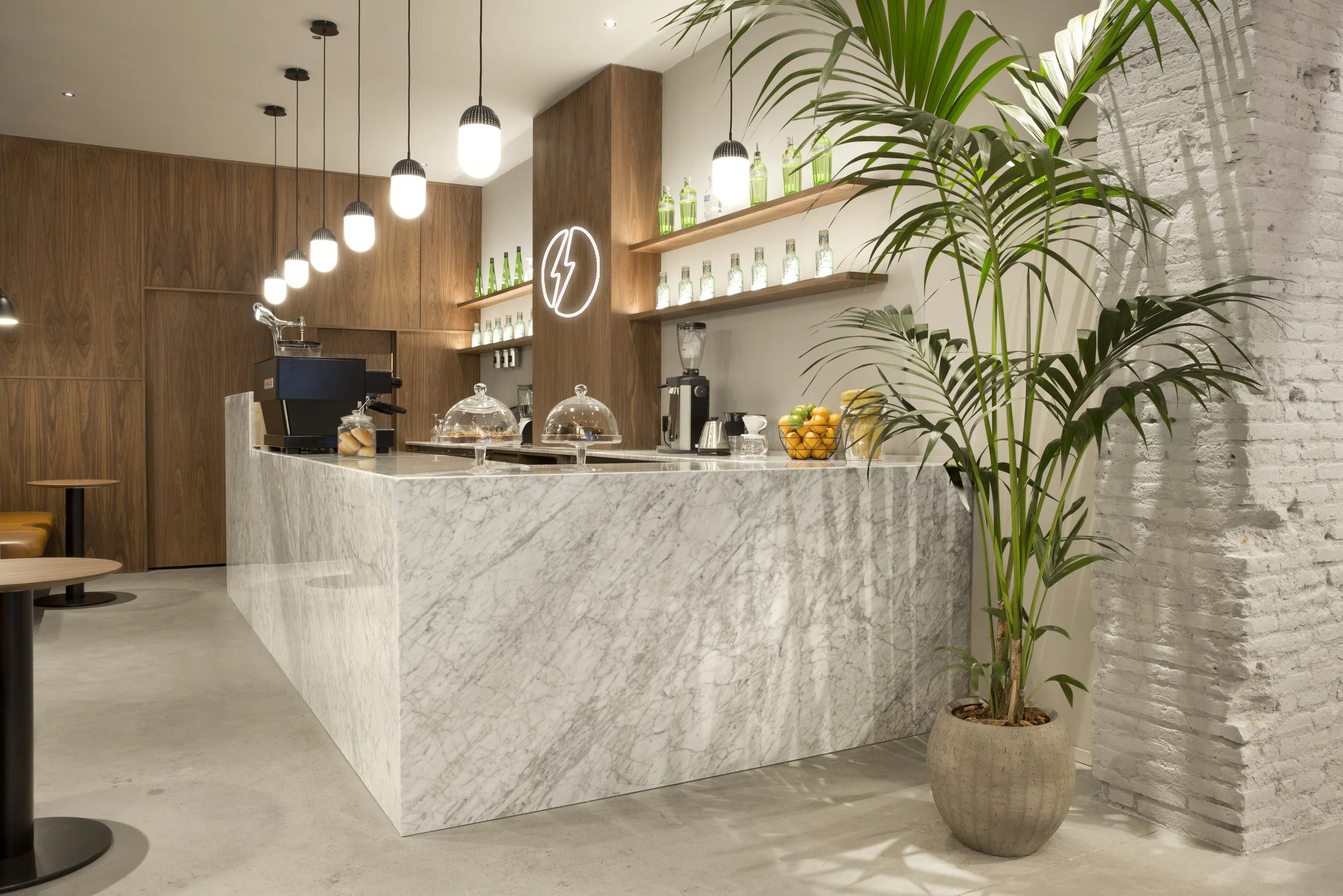 Interiores de cafetería con una barra de mármol blanco, estanterías con botellas verdes, lámparas colgantes y plantas en macetas, paredes de ladrillo pintado de blanco.