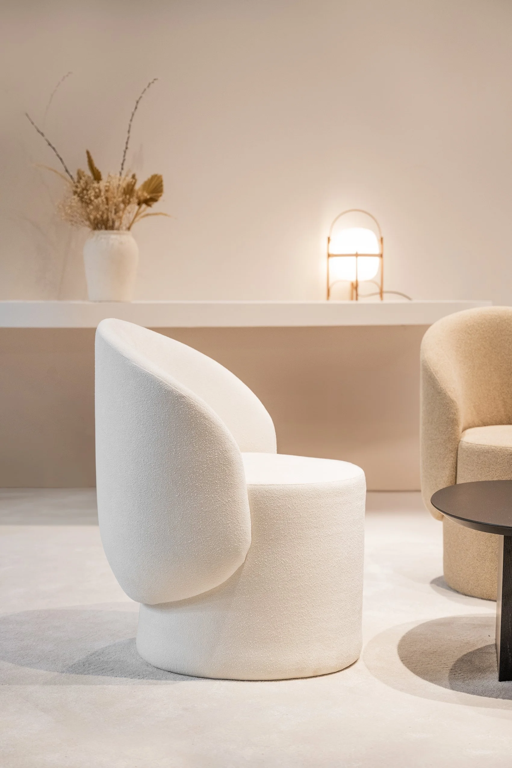 Silla de color blanco y suave con forma moderna en primer plano, otra silla beige y una mesa negra pequeña a un lado, en un espacio minimalista con pared clara, una lámpara y un jarrón con flores secas en una repisa.