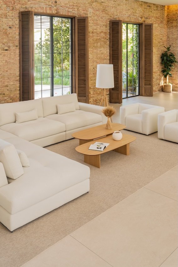 Sala de estar con paredes de ladrillo, grandes ventanas con persianas de madera, sofá y sillones blancos, mesa de centro de madera con decoraciones, lámpara de pie blanca, planta en una maceta y vista a un espacio exterior con árboles.