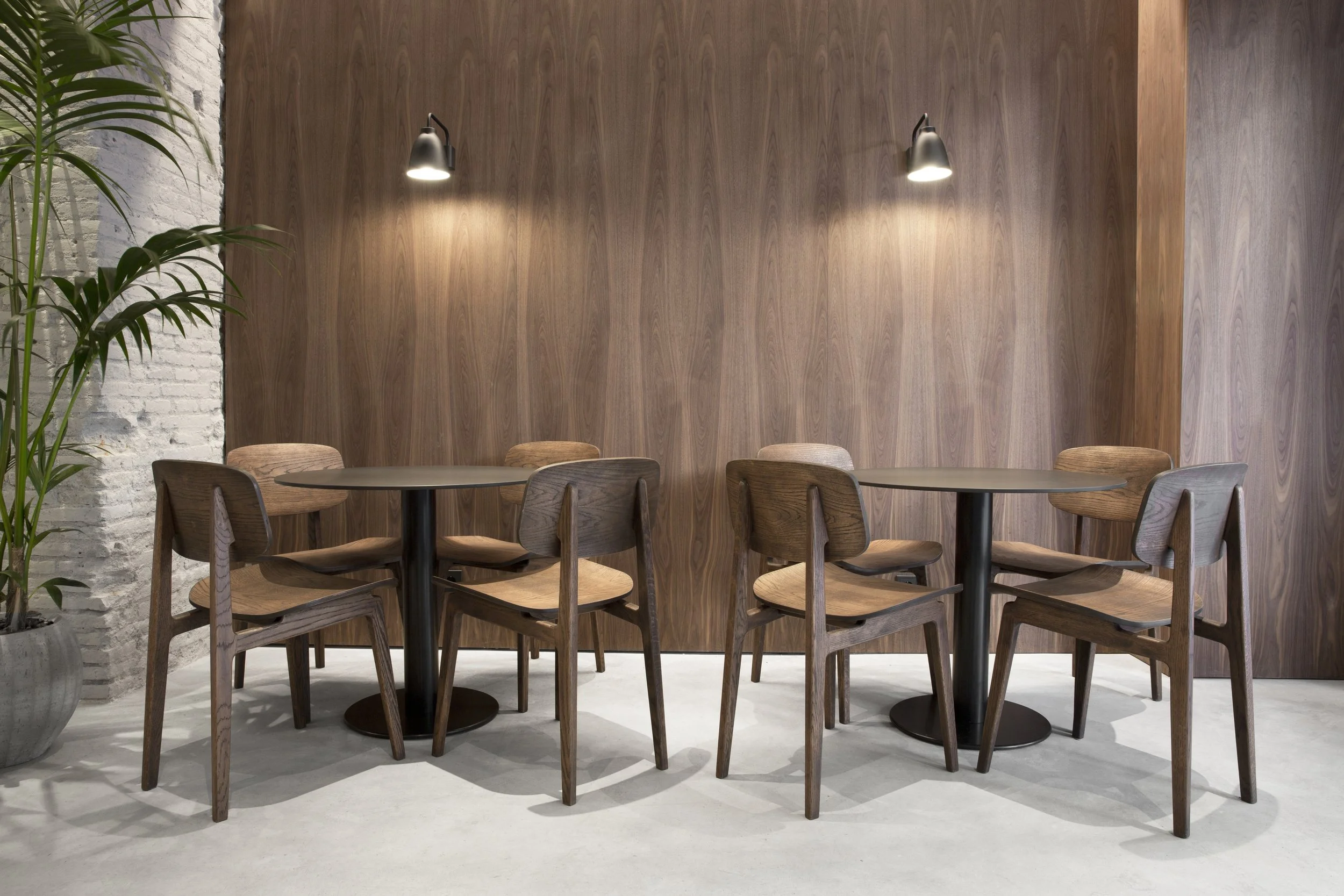 Sala moderna con paredes de madera, lámparas de pared, mesas redondas y sillas de madera, planta en receptáculo de cemento.
