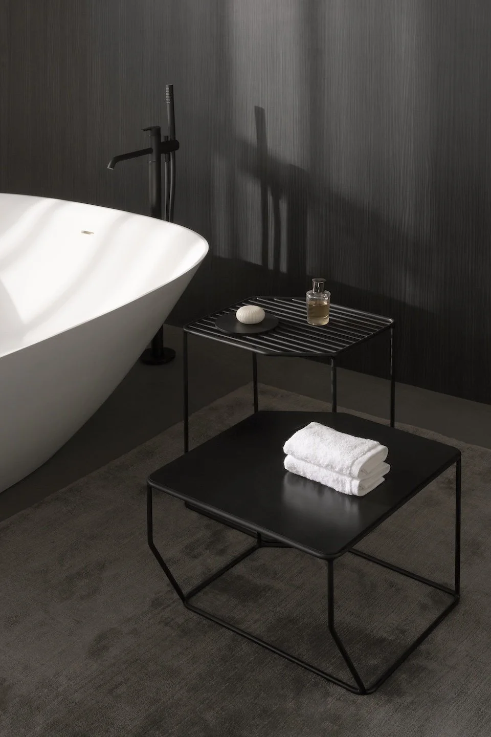 Una bañera de diseño moderno en un baño de estilo minimalista con paredes negras oscuras, un mueble auxiliar negro y un conjunto de toallas blancas dobladas.