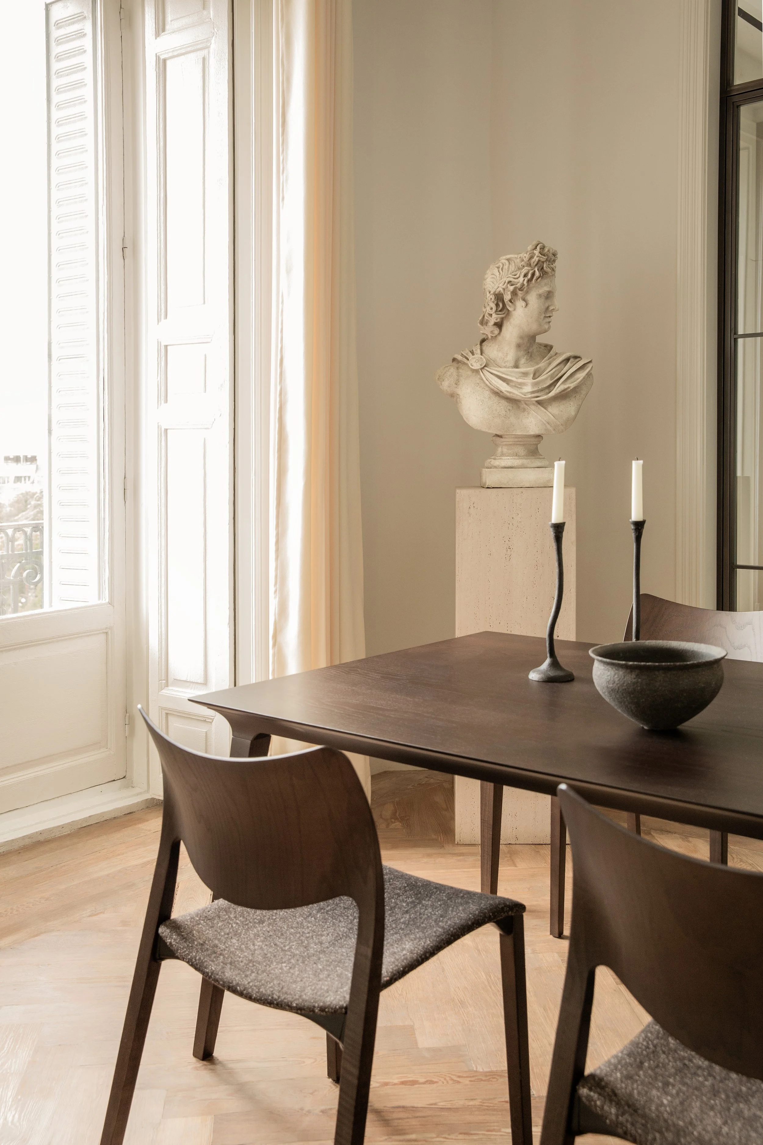 Interior de una sala con una mesa de madera oscura rodeada de sillas de madera con asientos de tela gris. En el fondo, una escultura de busto de una figura clásica, sobre un pedestal de piedra, y dos candelabros con velas en la mesa. Frente a una ventana grande con cortinas claras, paredes blancas y una vista parcial de un balcón.