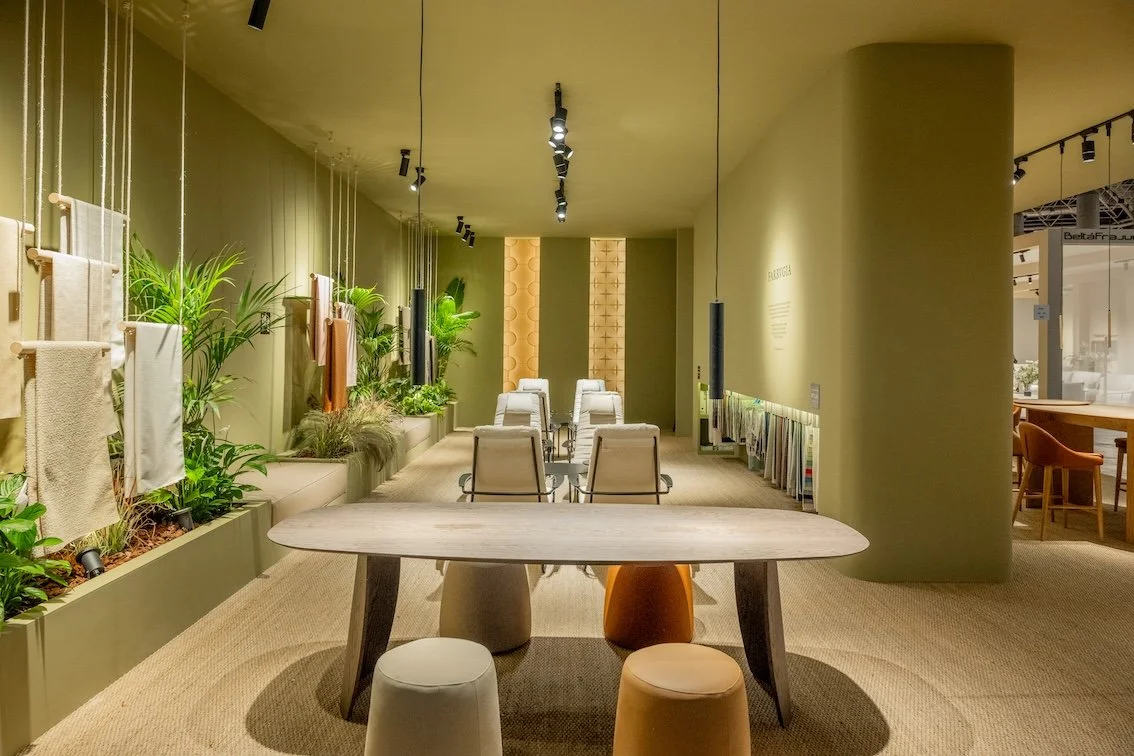 Espacio interior con ambiente natural, plantas y telas decorativas en las paredes, sillas y mesa en el centro, iluminación moderna y paredes en tonos tierra.