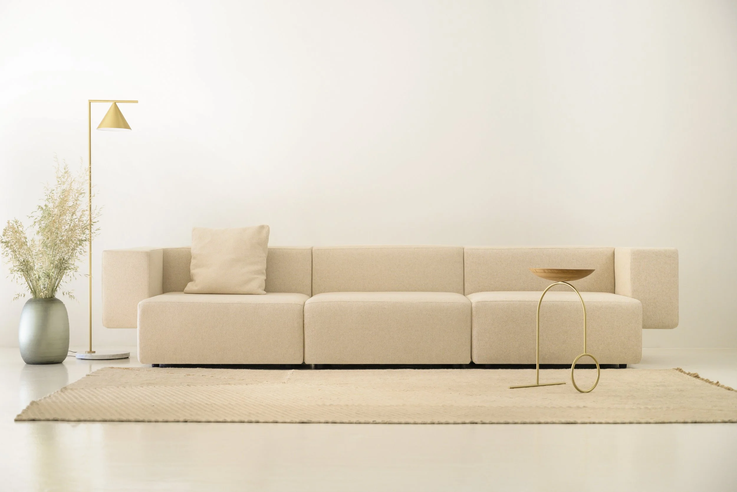 Sala de estar minimalista con sofá beige, alfombra en color crema, lámpara de piso dorada, jarrón con plantas y mesa decorativa en tonos dorados.