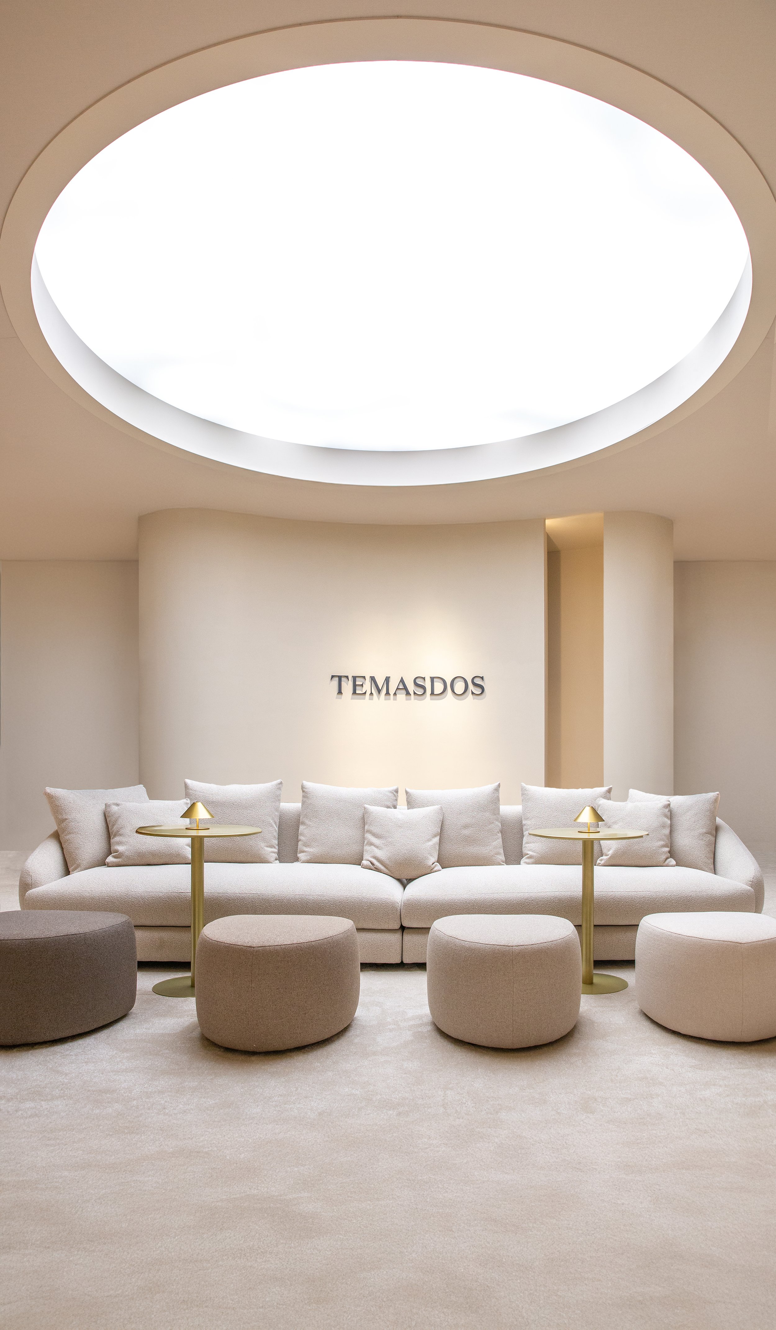 Sala de estar con sillones blancos, mesitas pequeñas con lámparas doradas, tapete beige y pared con la palabra 'TEMASDOS' en letras negras. Techo con iluminación circular.