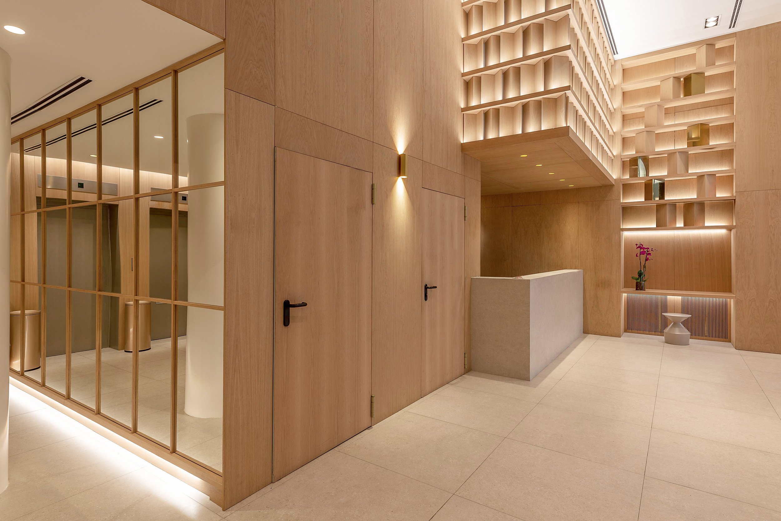Vestíbulo moderno con paredes y techos de madera clara, iluminación empotrada y decoraciones minimalistas, incluyendo una planta de orquídea en una mesa de recepción.