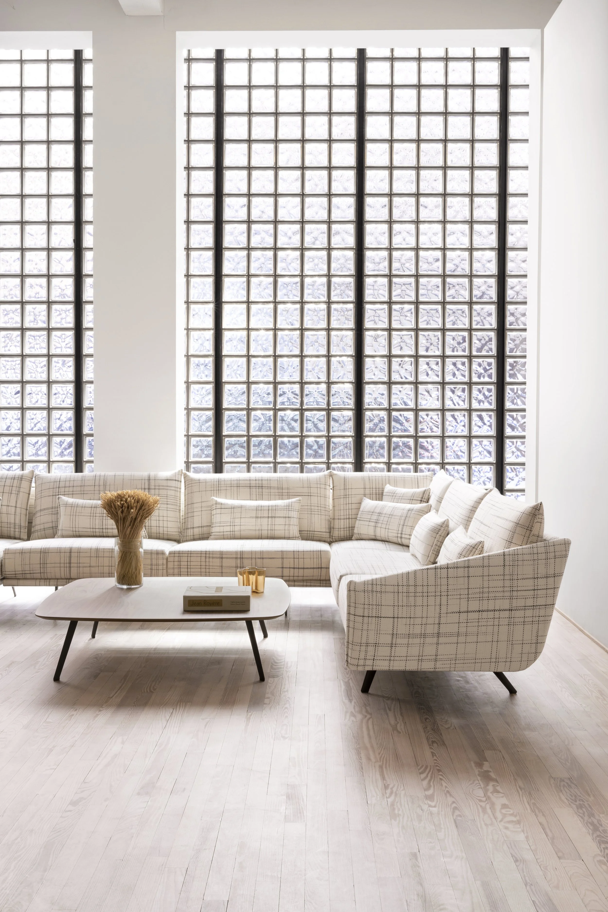 Sala moderna con una gran ventana de bloques de cristal, sofá beige con cojines a cuadros, mesa de centro blanca con libros y decoración, suelo de madera clara.