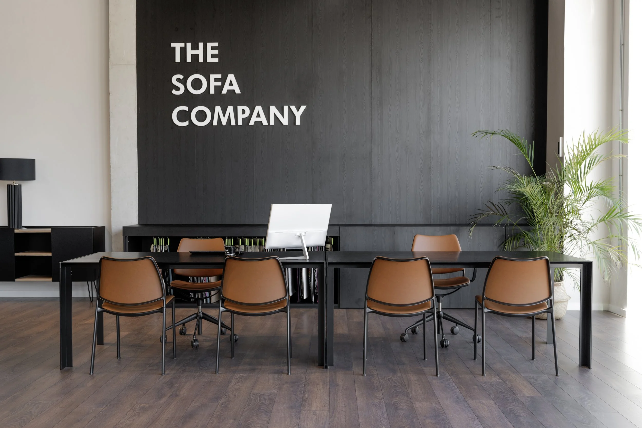 Sala de reuniones moderna con una mesa negra y sillas de color marrón, pared de fondo gris oscuro con letras blancas que dicen 'The Sofa Company', interior iluminado por luz natural y plantas decorativas.