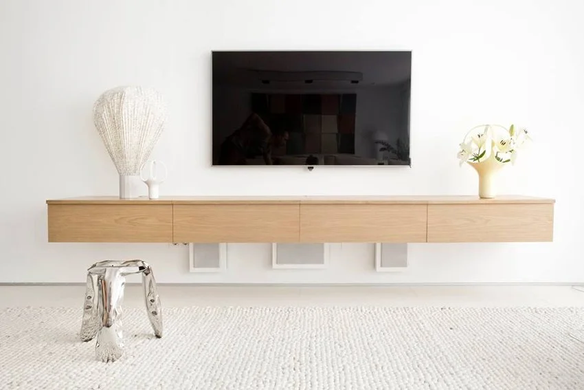 Sala minimalista con pared blanca, televisor negro montado y mueble de madera, decorado con jarrones de cerámica y flores blancas, con silla metálica moderna y alfombra blanca.