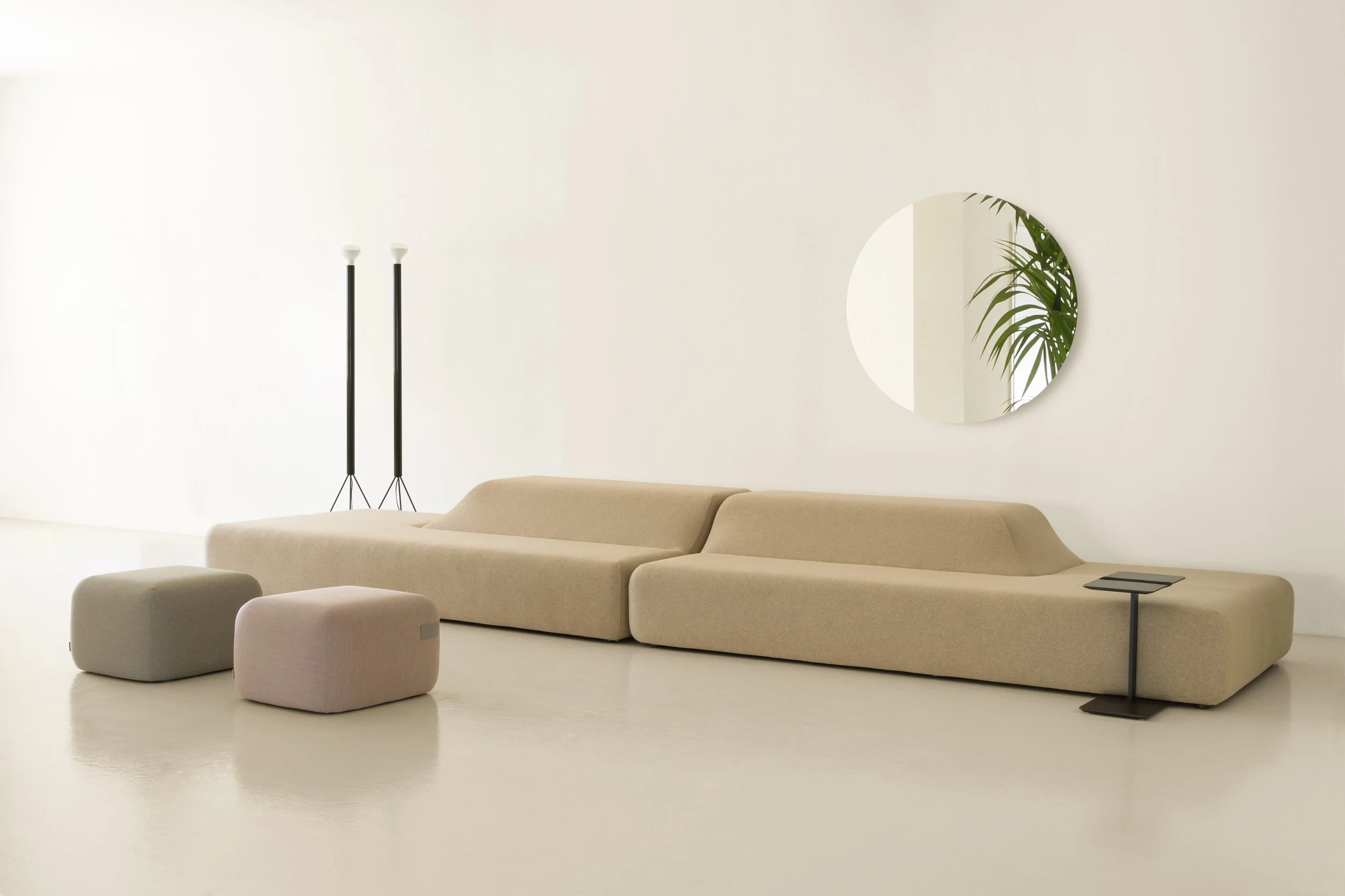 Sala moderna con sofá beige, ottomans del mismo color, espejo redondo en la pared y lámparas de pie negras con luces blancas, piso blanco y paredes blancas.
