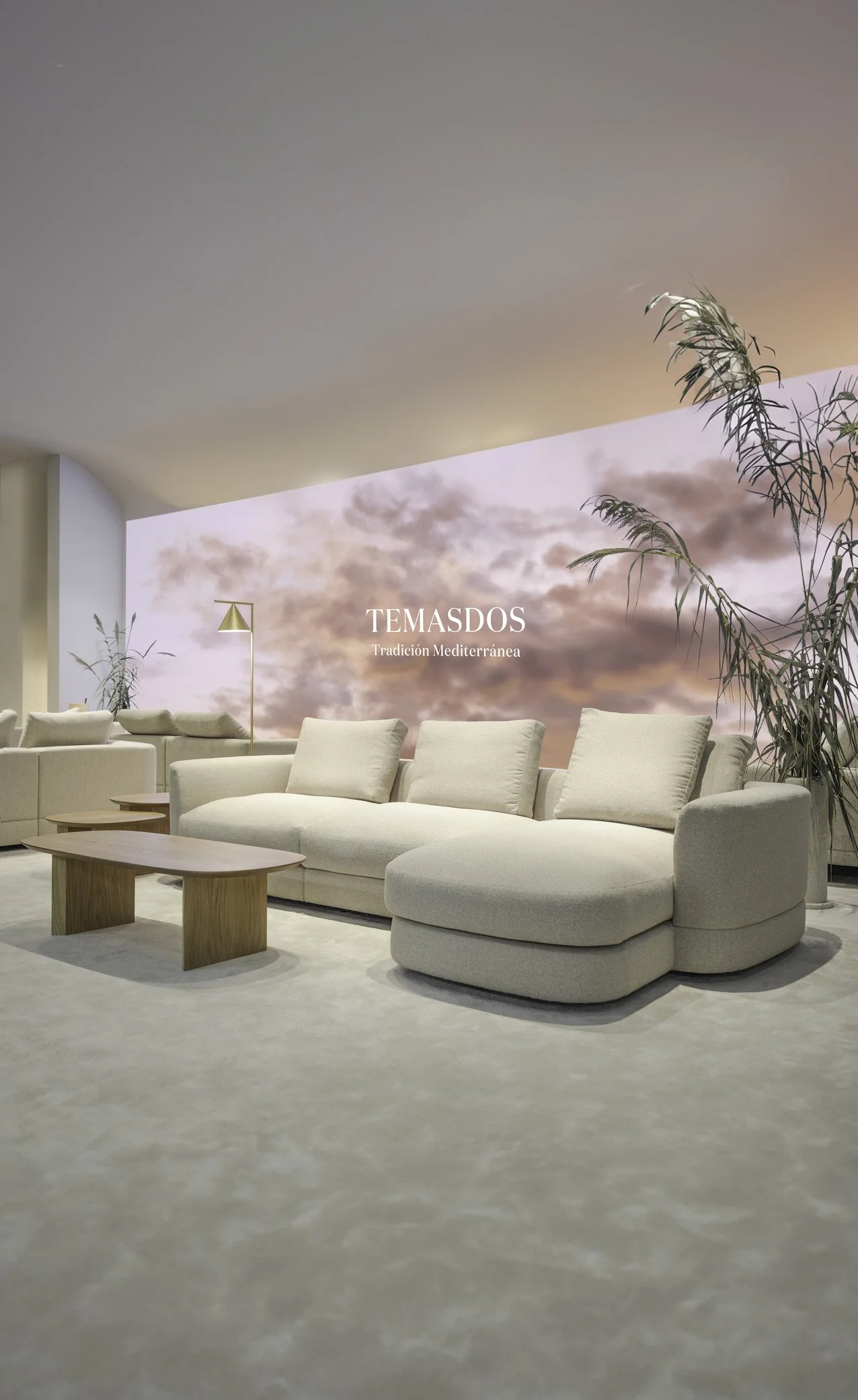Sala de estar moderna con sillones blancos, mesas de madera, lámpara dorada y plantas, con un mural de cielo al atardecer y texto que dice 'TEMASDOS, Tradición Mediterránea'