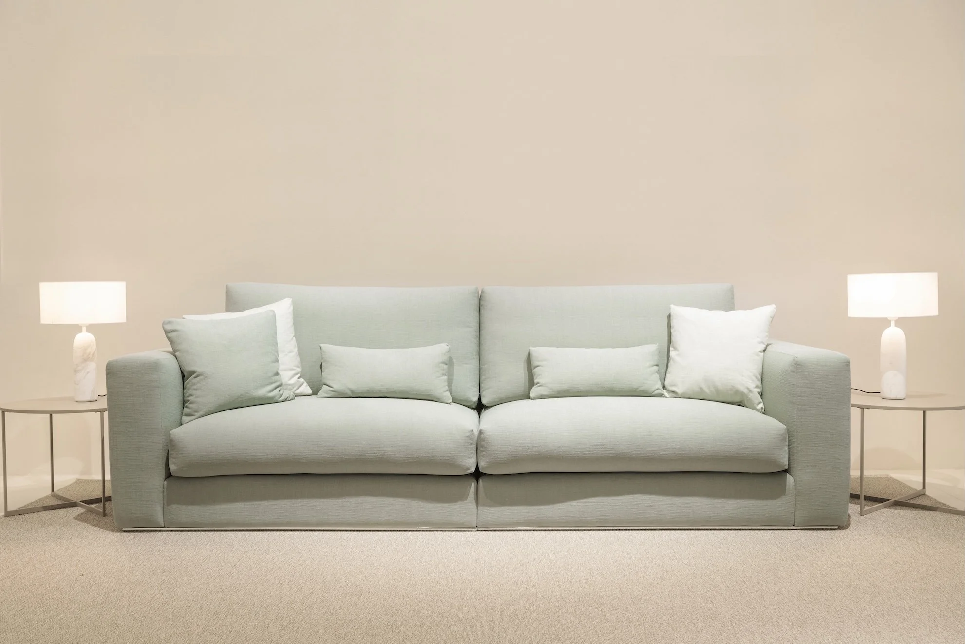 Sofá gris claro con cinco cojines en una sala minimalista, con lámparas blancas a cada lado y muro beige de fondo.