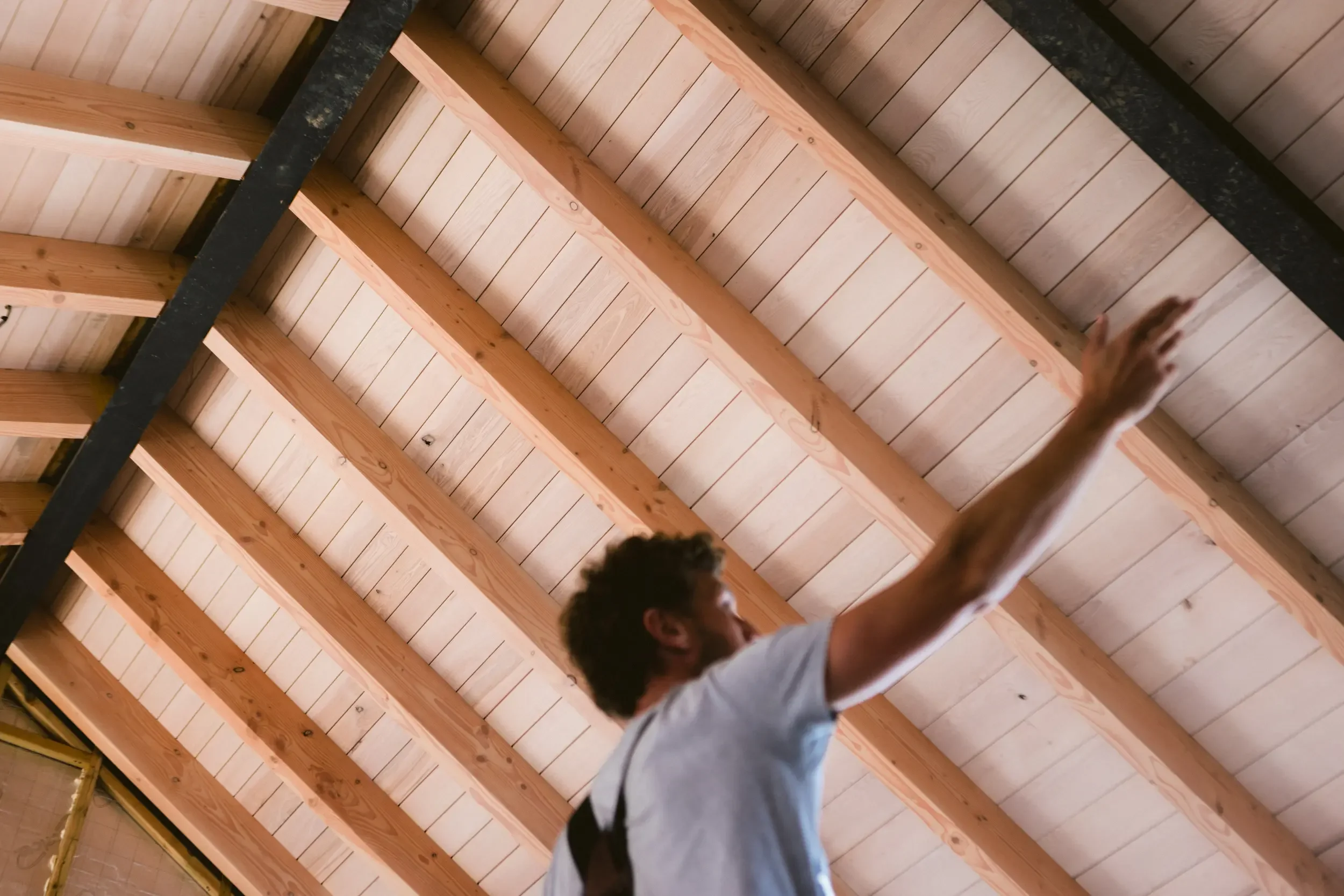Man reaching up with one arm towards a wooden ceiling with exposed beams.