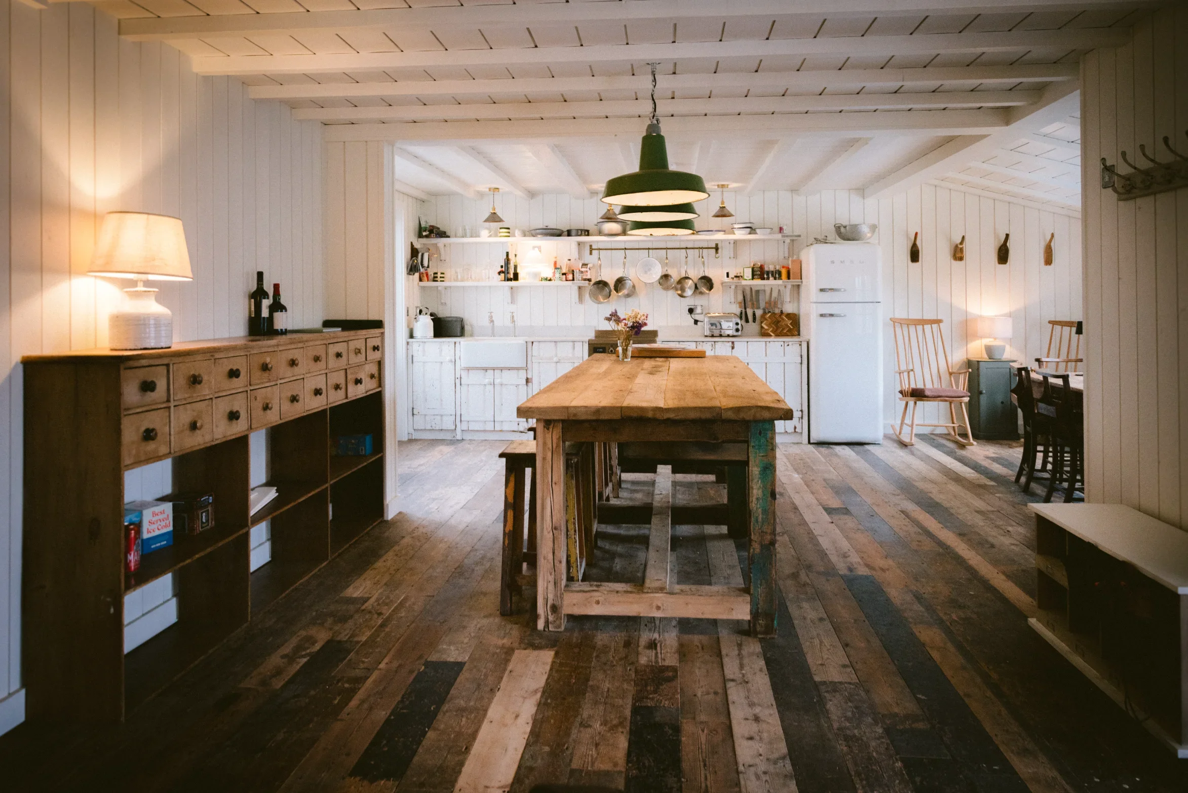 Open-plan kitchen and dining room with reclaimed timber floor, exposed painted beams and full-width shelving, Cornwall conversion