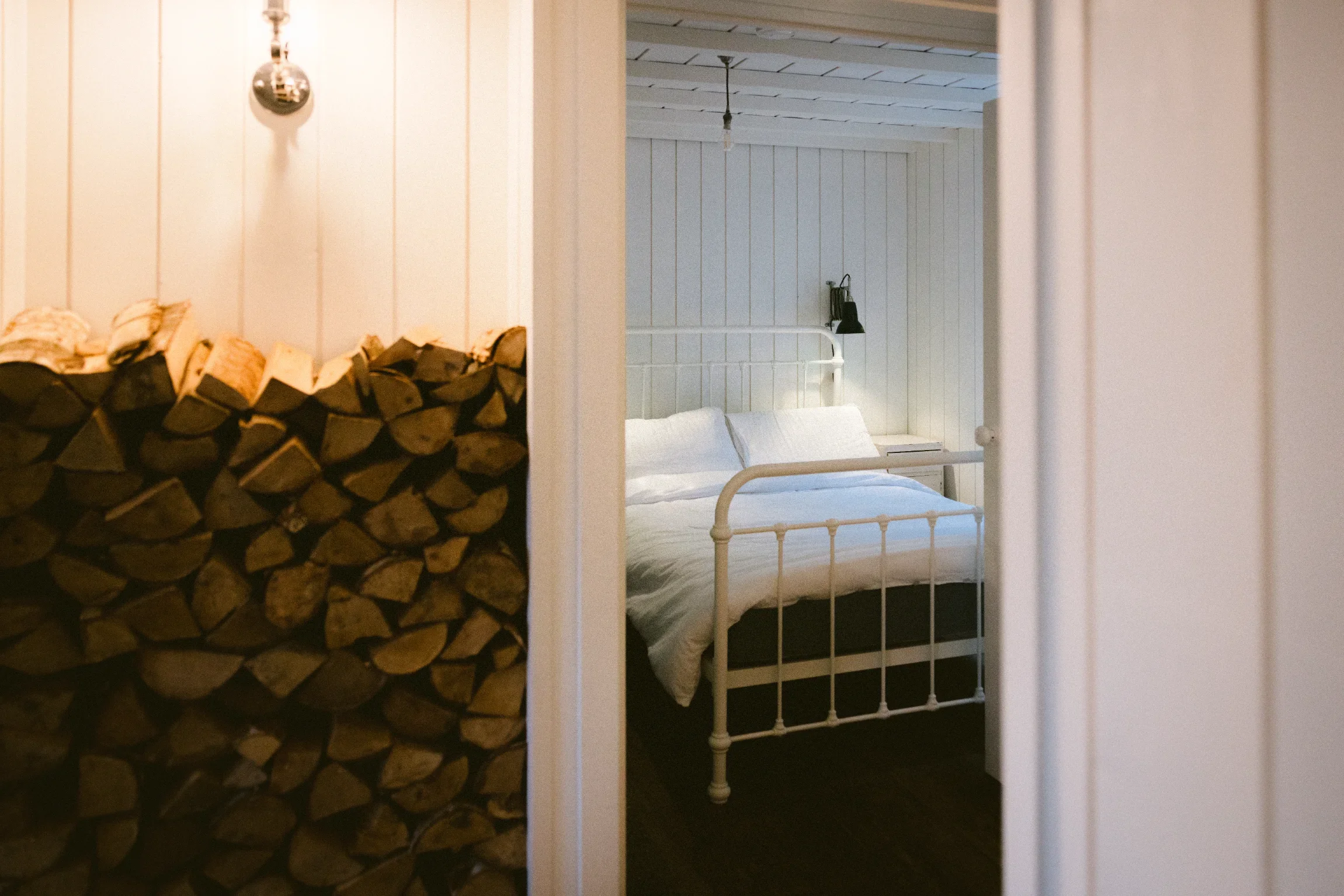Bedroom interior glimpsed through doorway alongside indoor log store, tongue-and-groove lined walls, Chapel Porth conversion Cornwall