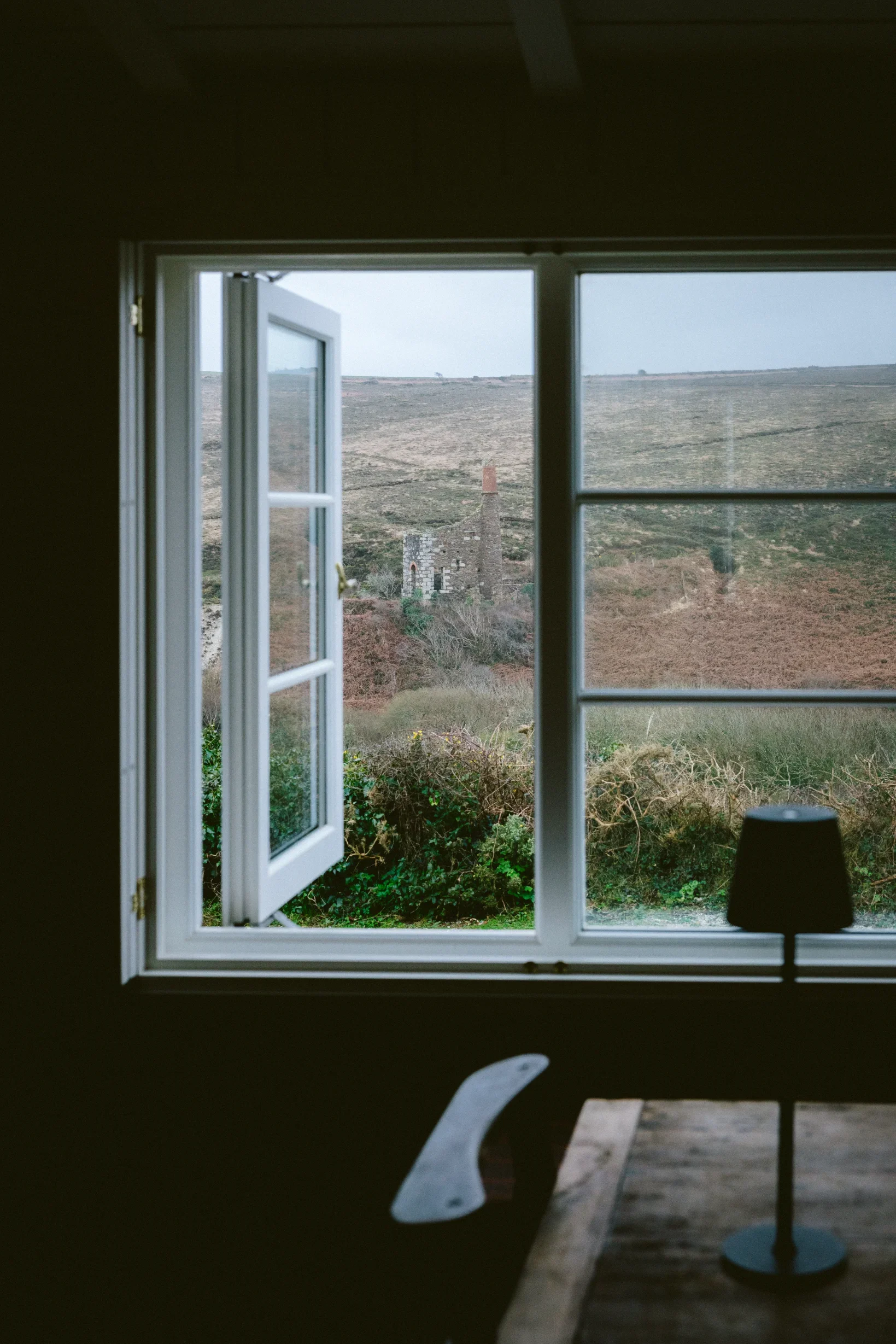 Open casement window framing a derelict Cornish tin mine engine house across moorland, Chapel Porth, St Agnes