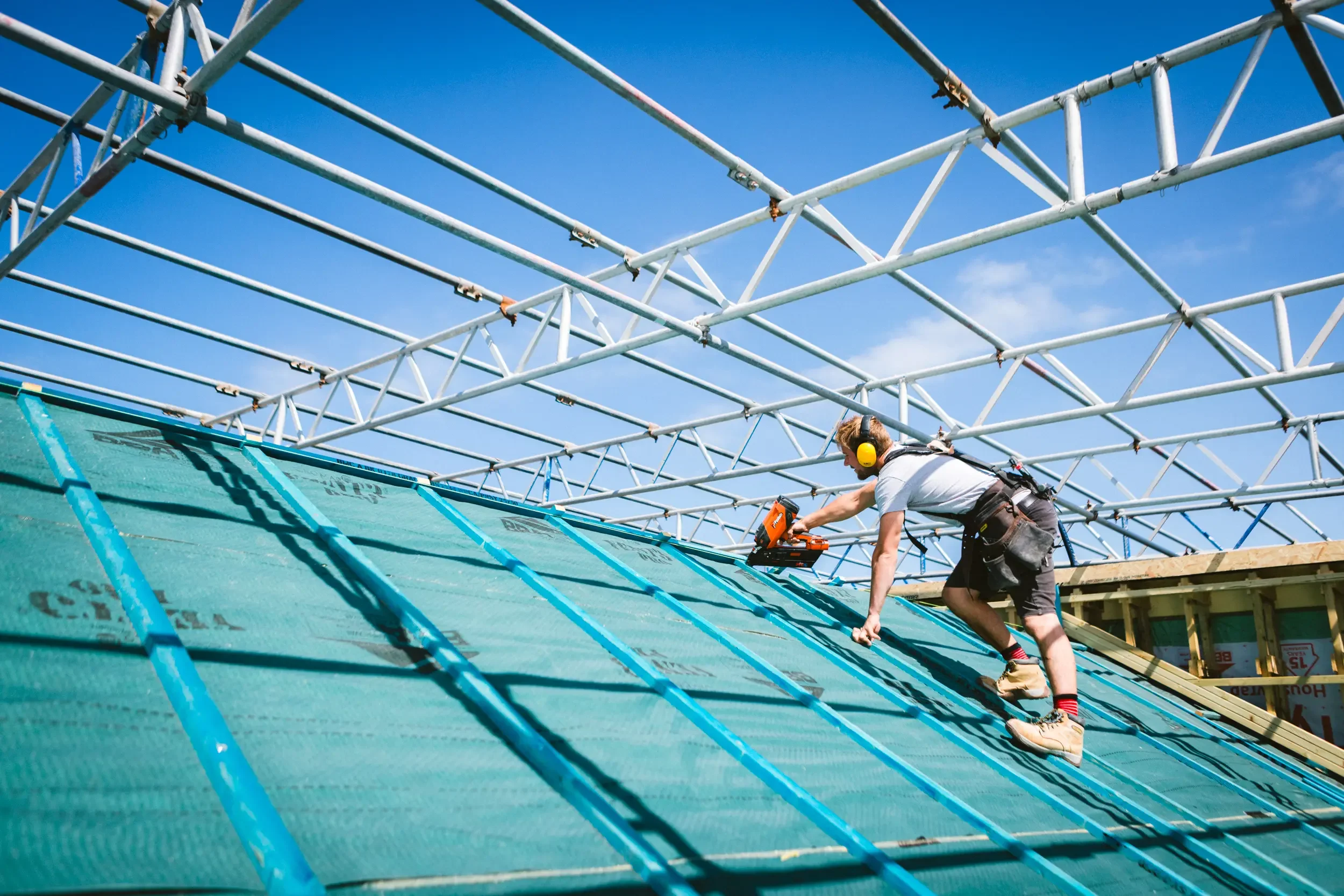 A worker wearing a white shirt, shorts, and yellow ear protection is installing roofing materials on a sloped roof, using a power tool, with metal scaffolding overhead and a clear blue sky in the background.