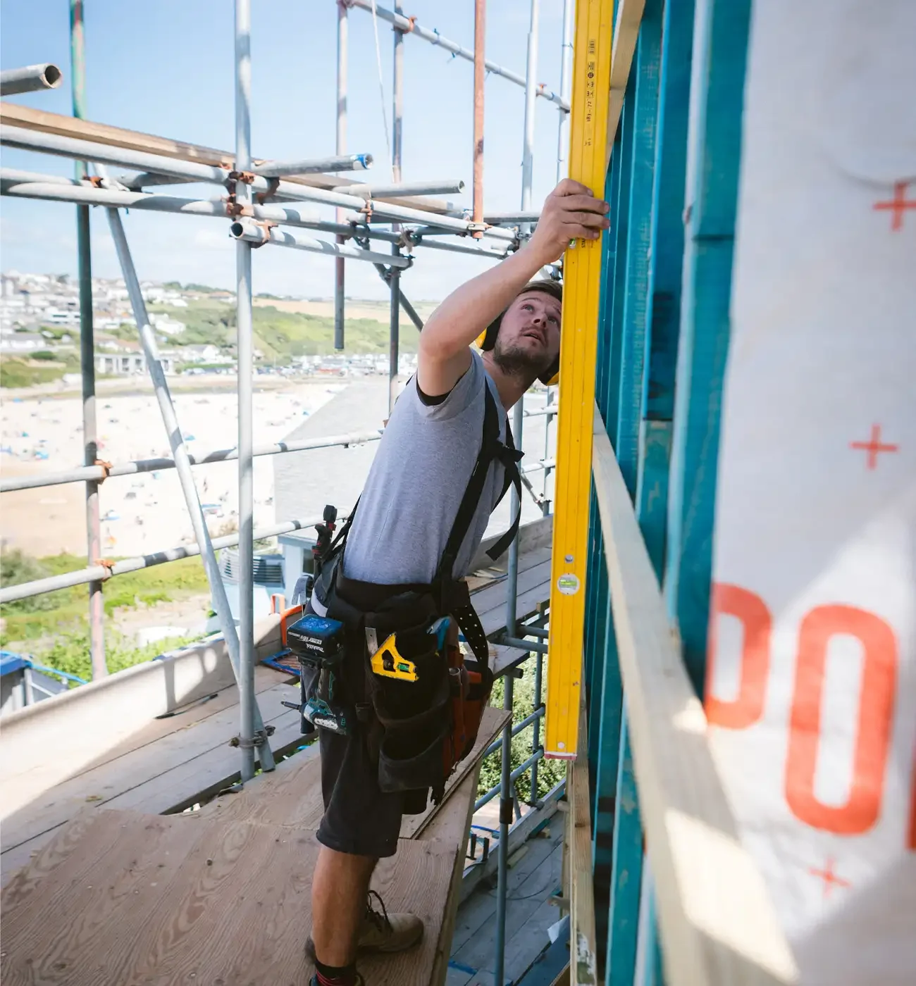 Elements Build carpenter checking vertical level on timber frame wall using a spirit level, scaffolding overlooking a Cornwall beach