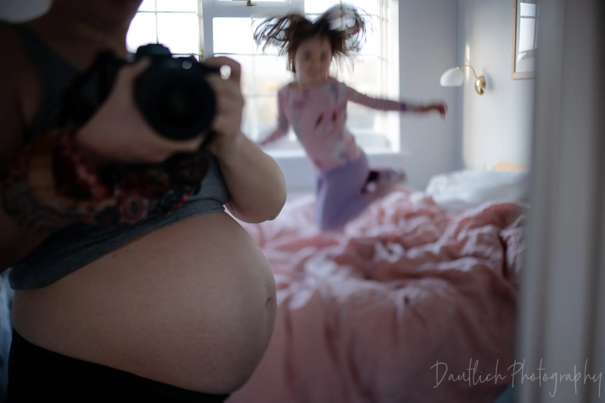 toddler jumping on bed with crazy hair while pregnant mother takes a photo in the mirror