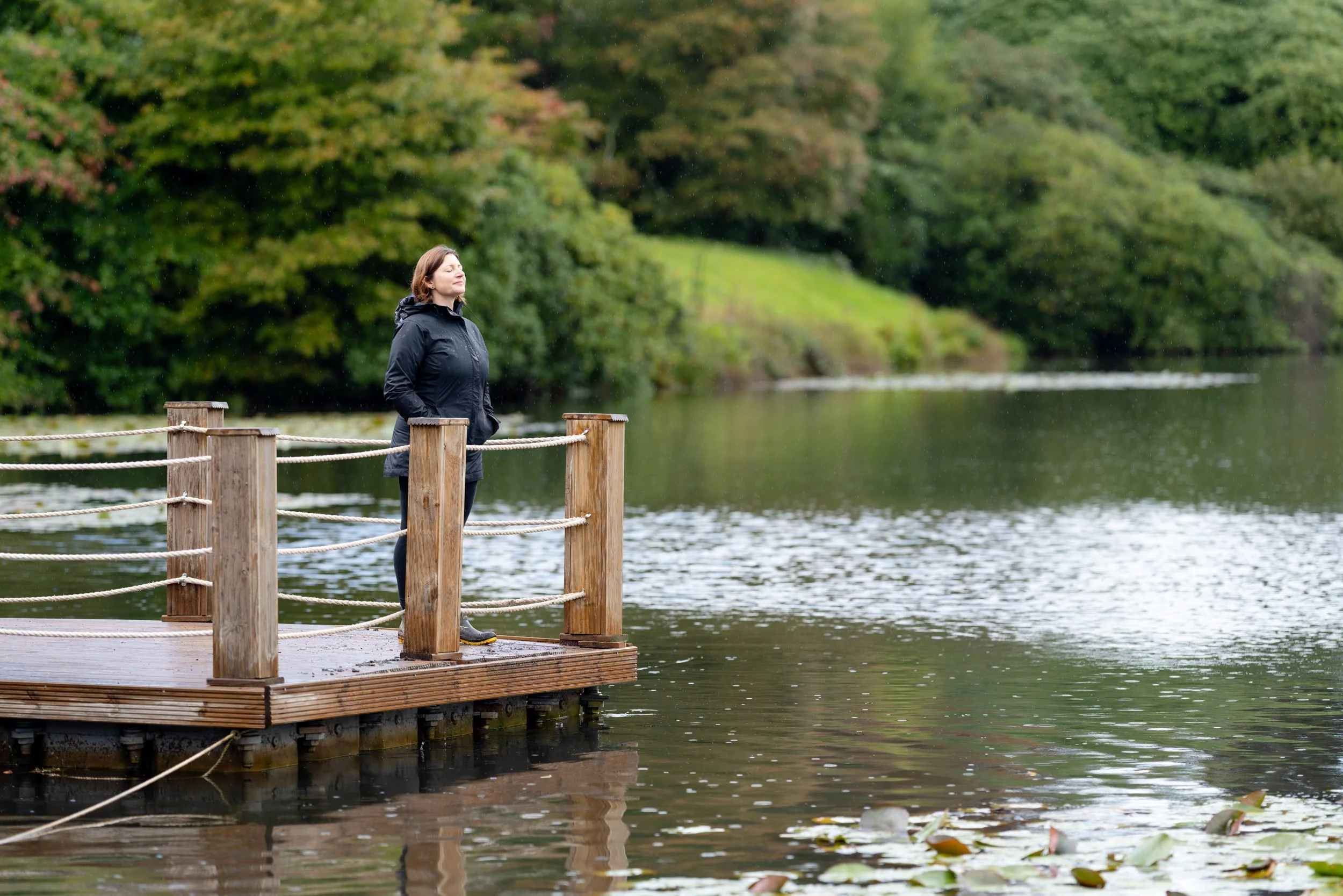 A woman standing on a wooden dock by the water, with trees in the background and rain falling.
