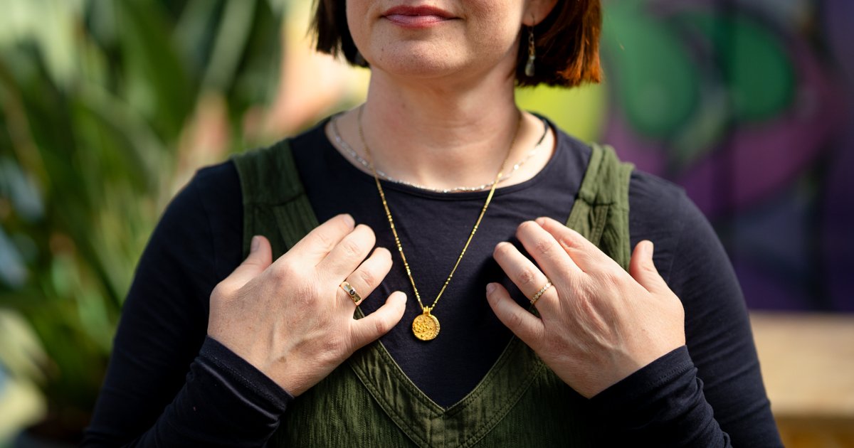 Woman tapping on her collarbone meridian point wearing a black shirt and green jumpsuit with a brightly coloured background