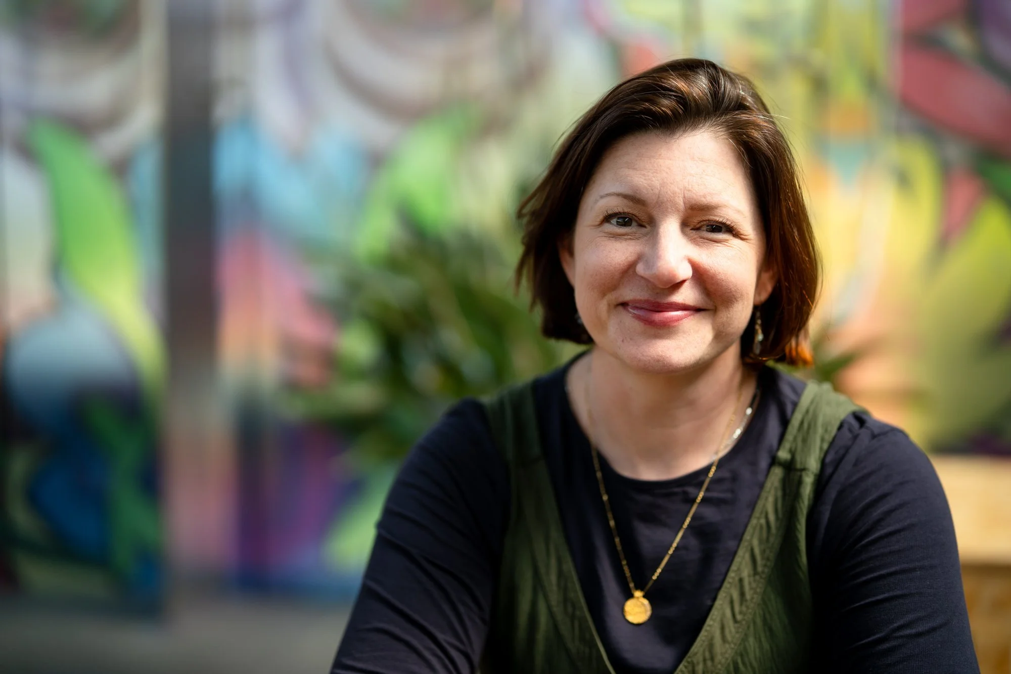 Woman looking at camera wearing a black long sleeve shirt and gold necklace with a bright wall behind her