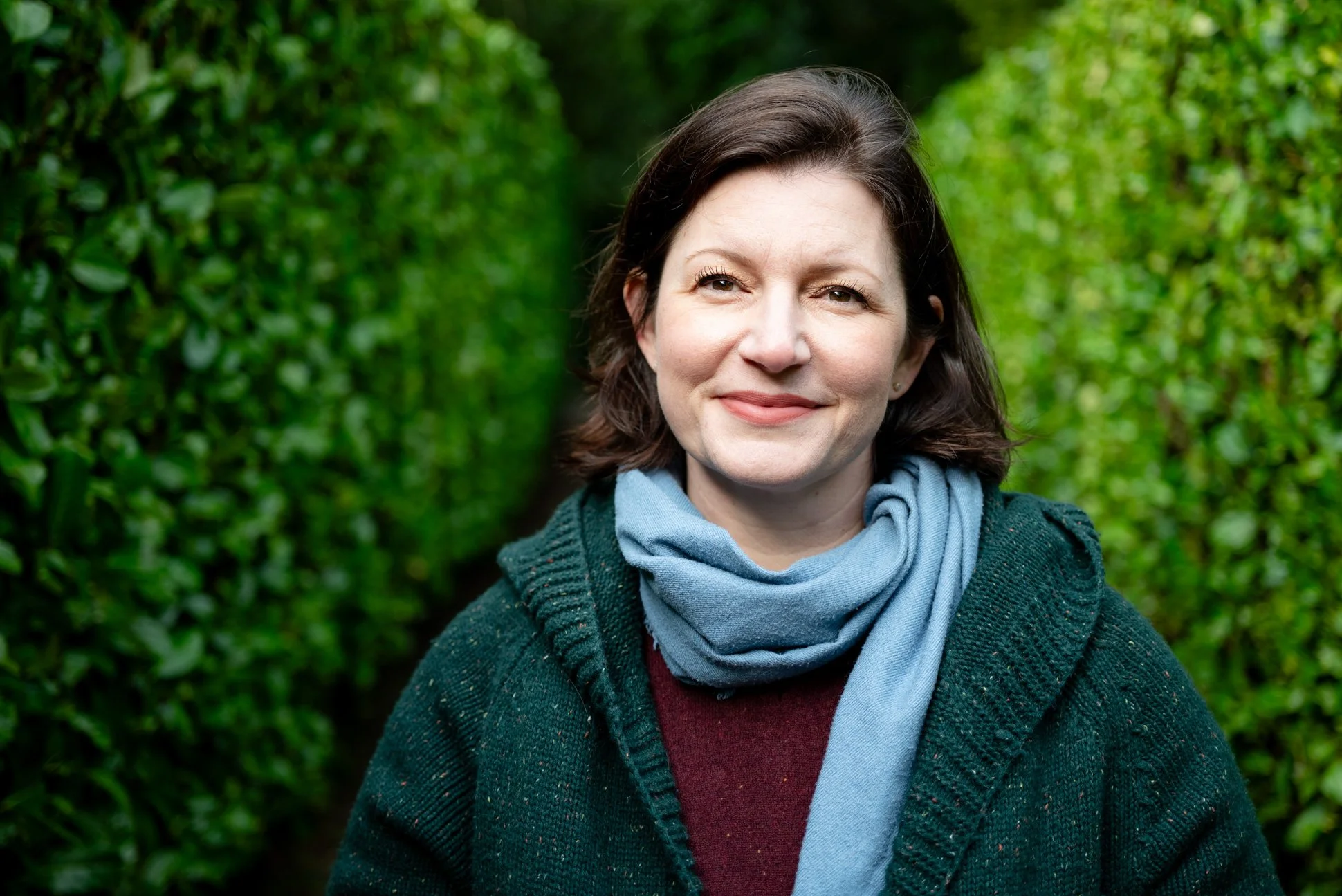 April Dautlich smiling while standing in a row of green hedges with a blue scarf around her neck and a green button up cardigan on.
