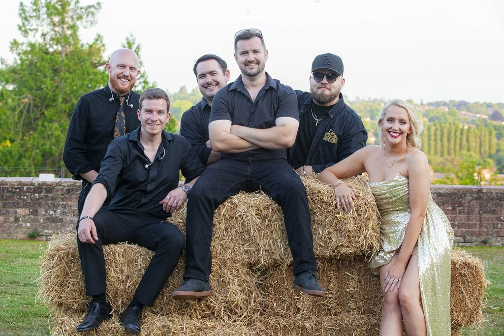 Group of six people posing outdoors on hay bales, with a brick wall and greenery in the background. Five men are dressed in dark clothing, with one woman in a shiny gold dress.