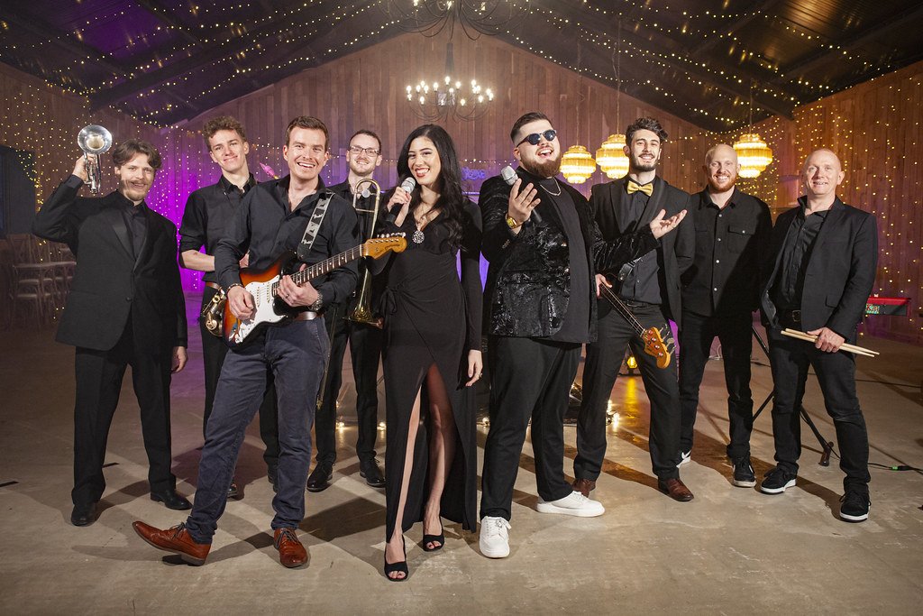 A diverse group of nine musicians and singers in formal black attire standing together in a decorated venue with string lights, chandeliers, and musical instruments.
