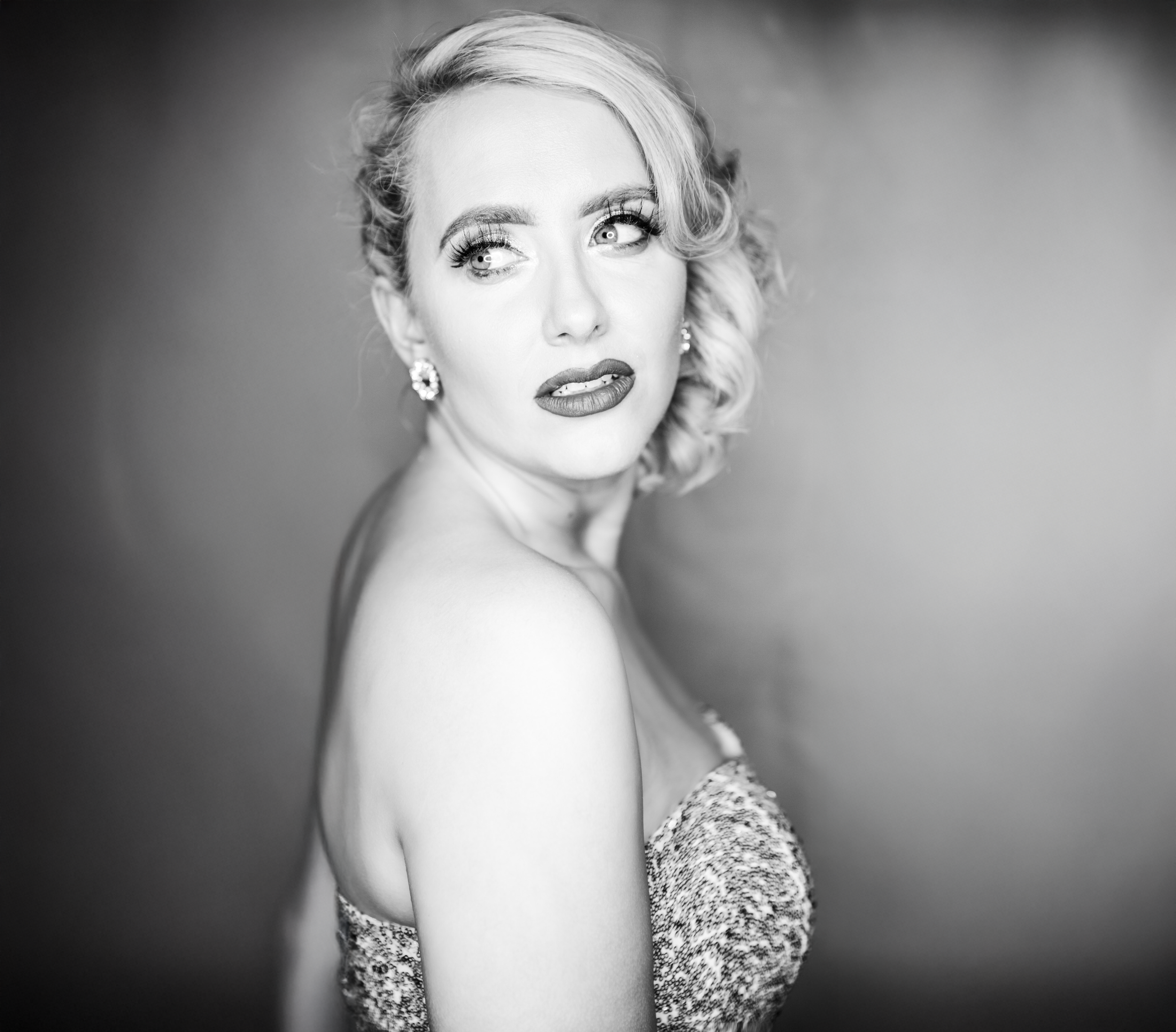 Black and white portrait of a woman with styled blonde hair, wearing earrings and a strapless dress, looking to her left with an expressive face.