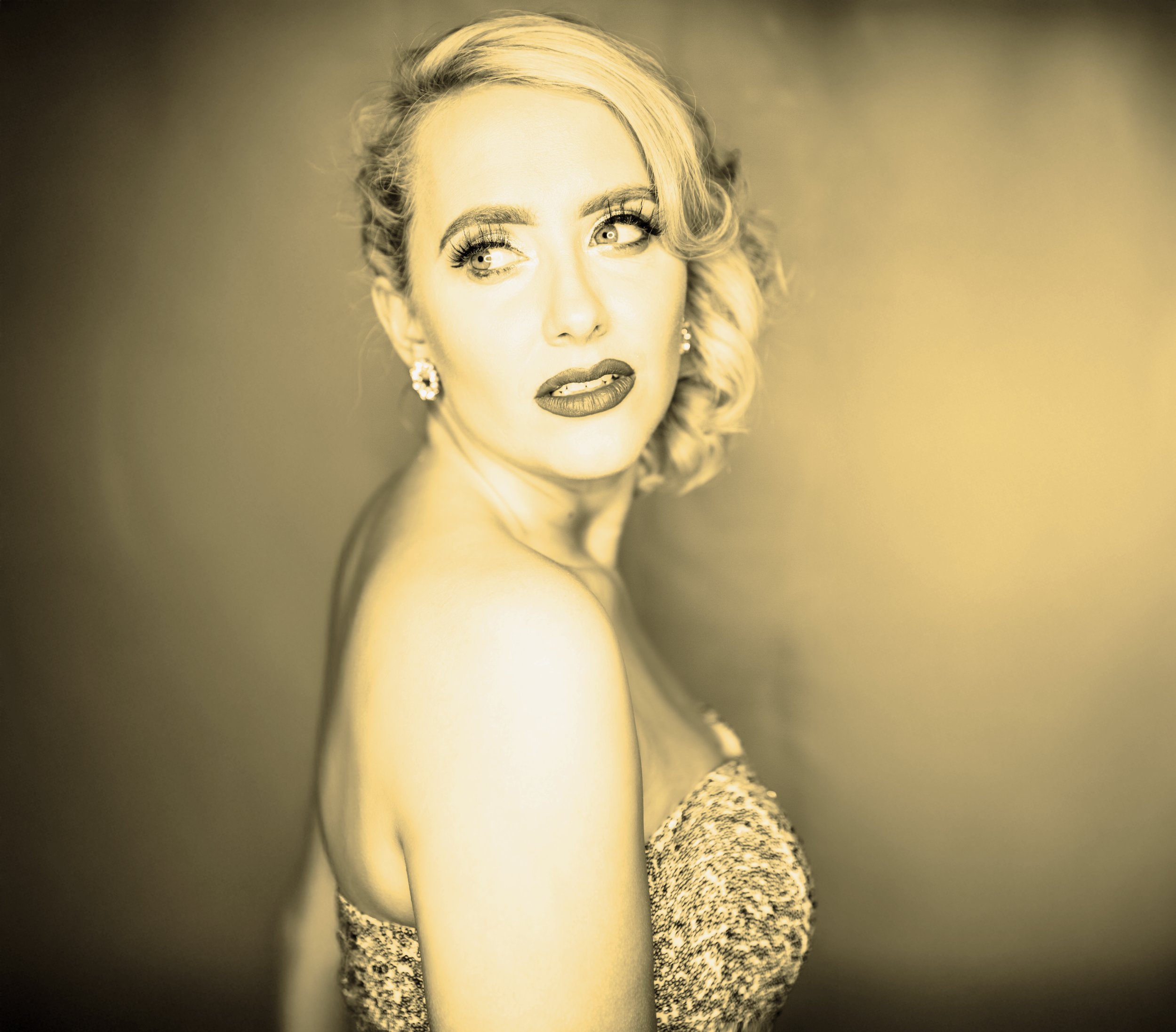 A woman with blonde, curly hair is looking over her shoulder. She is wearing earrings and a strapless, textured dress. The photo has a yellow tint with a dark background.