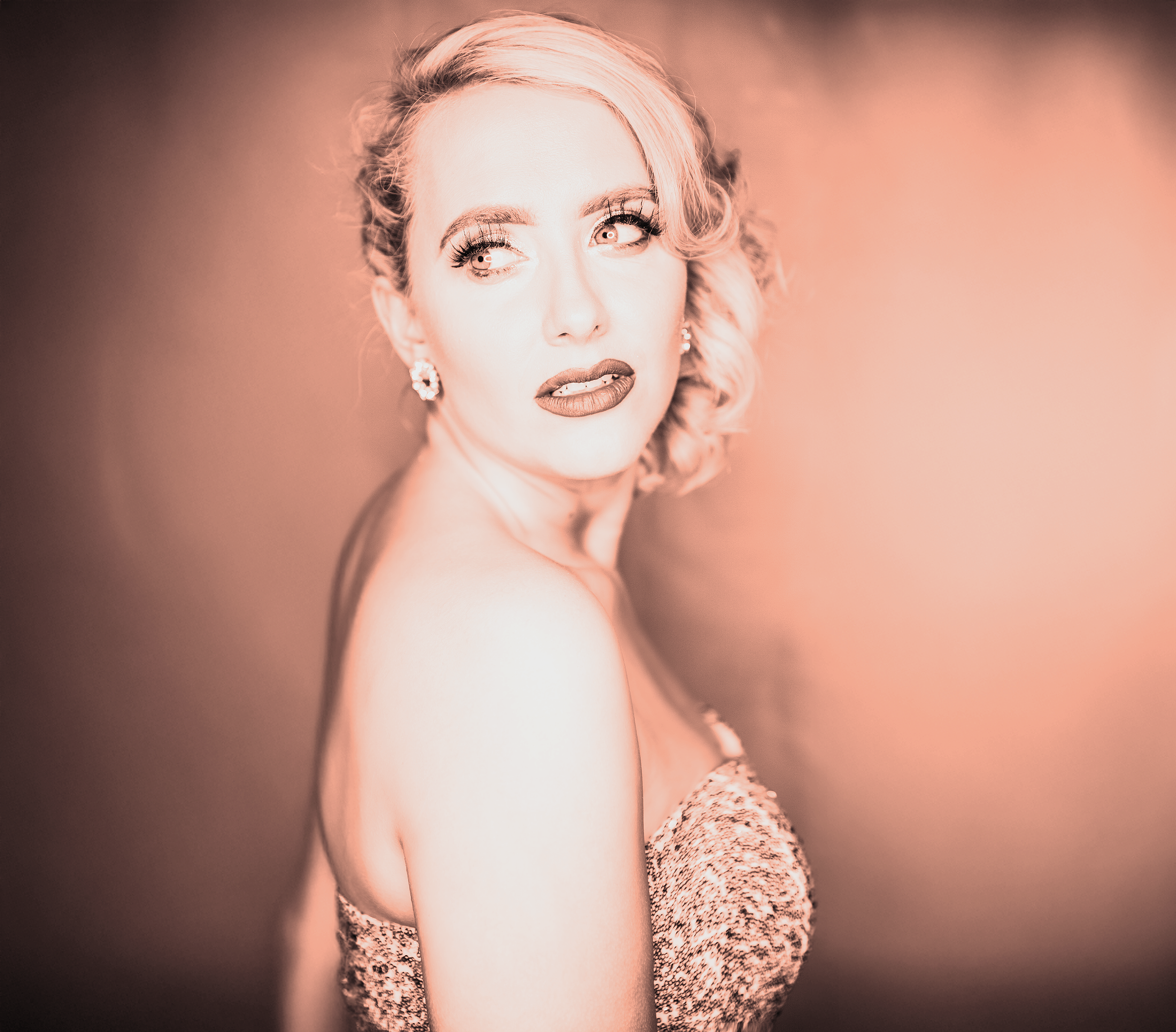 A woman with blonde hair styled in curls, wearing makeup with bold lipstick and earrings, poses with her body slightly turned, looking off to the side, against a gradient soft pink background.