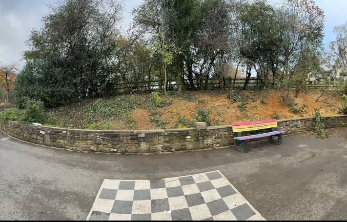 A colorful park bench with pink, yellow, purple, and black-painted slats situated next to a stone retaining wall, with a paved area and a chessboard pattern in front, surrounded by trees and shrubs on a cloudy day.