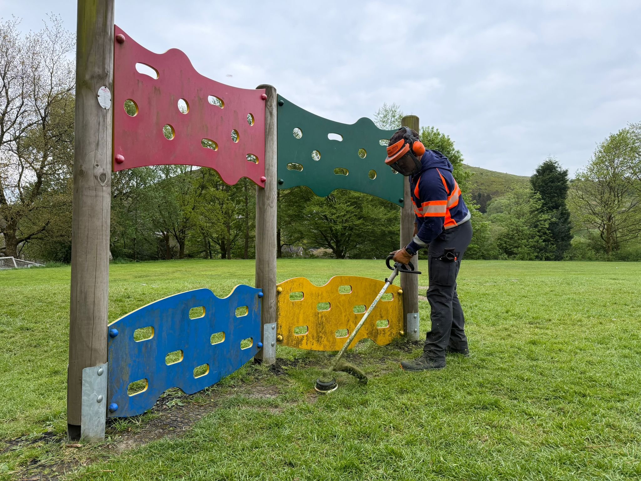 A worker wearing a safety helmet and vest is repairing or installing colorful outdoor playground panels with holes, set on wooden posts in a grassy park with trees and hills in the background.