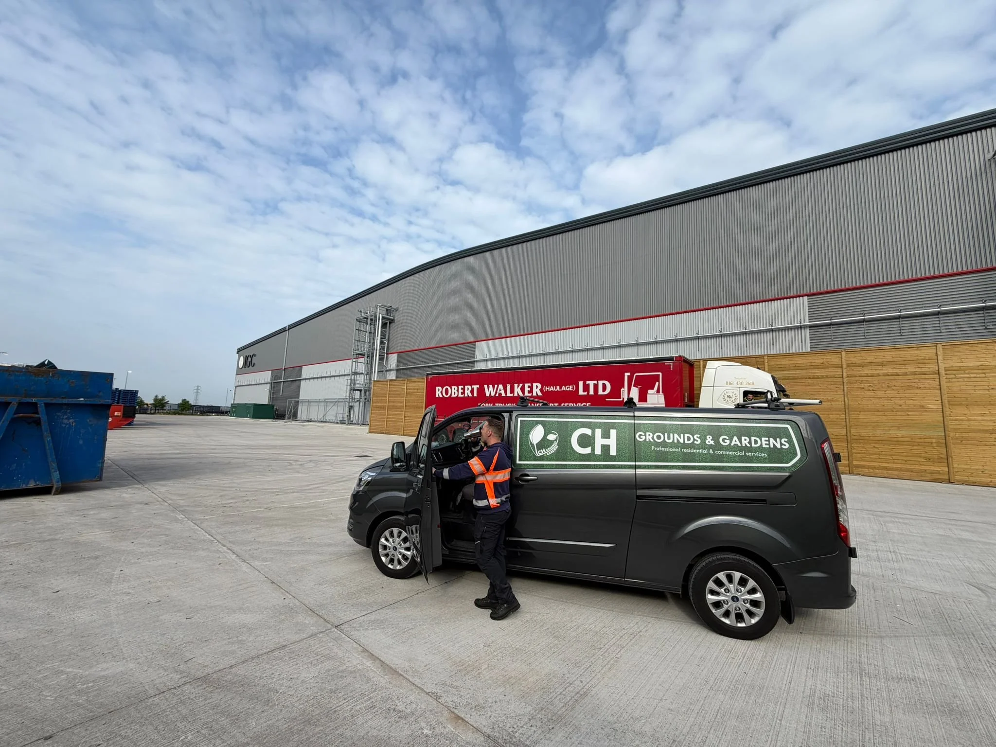 A worker standing outside a large industrial warehouse, opening the car door of a black vehicle. The warehouse has a curved gray metal exterior and a red sign that reads "ROBERT WALKER LTD." The sky above is partly cloudy.